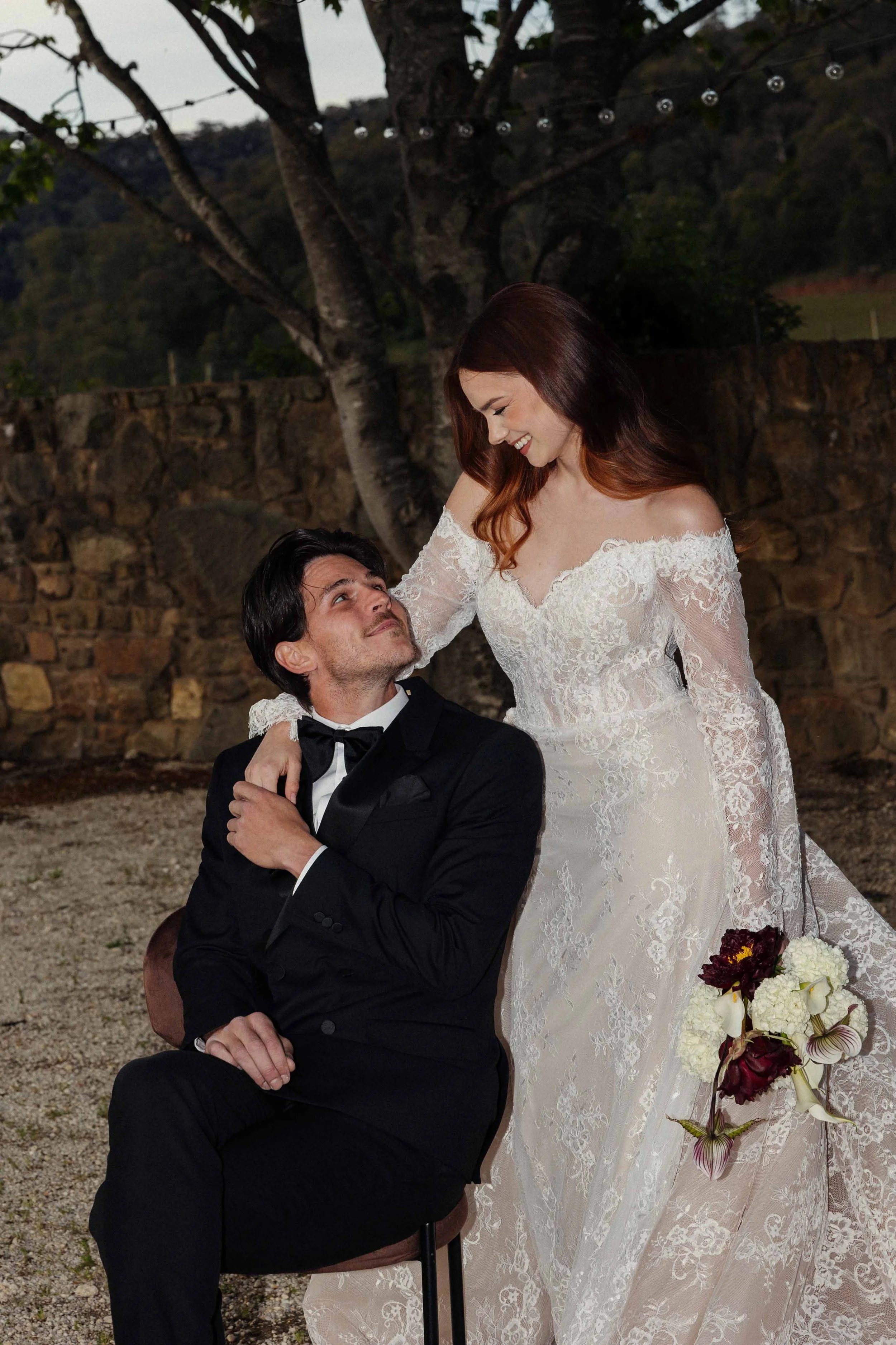 A bride in a lace wedding gown standing beside a groom in a black tuxedo, outdoors during evening with trees and string lights in the background.