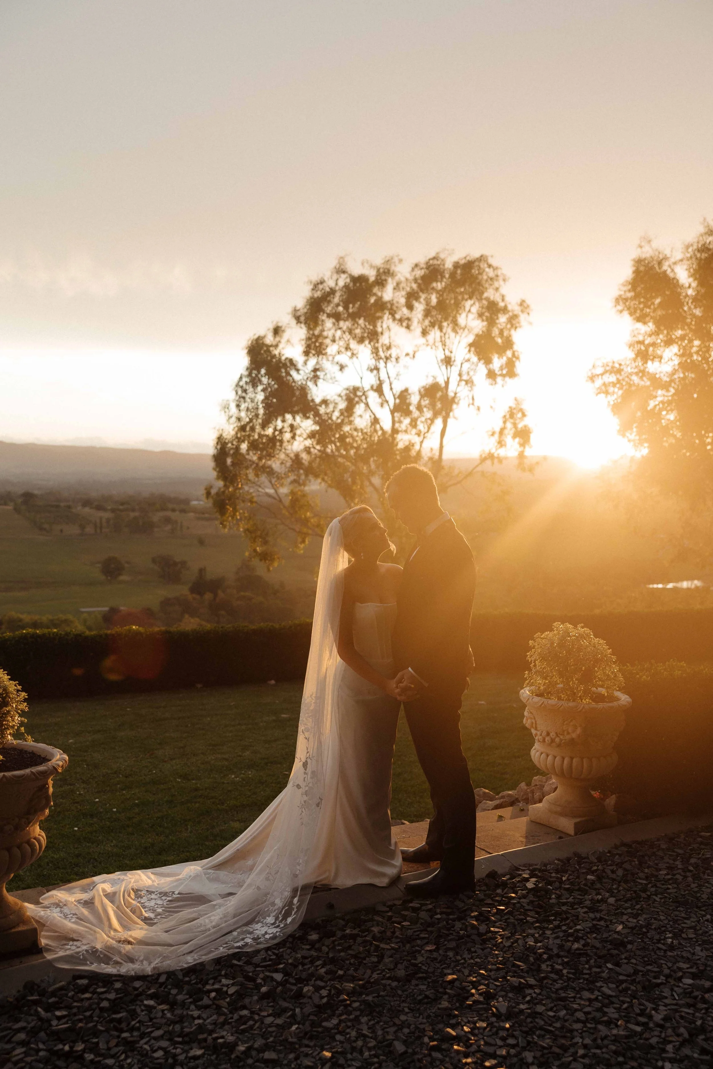 A bride and groom stand holding hands outdoors during sunset, with warm sunlight illuminating their silhouettes, in a scenic landscape setting with trees and pot planters.