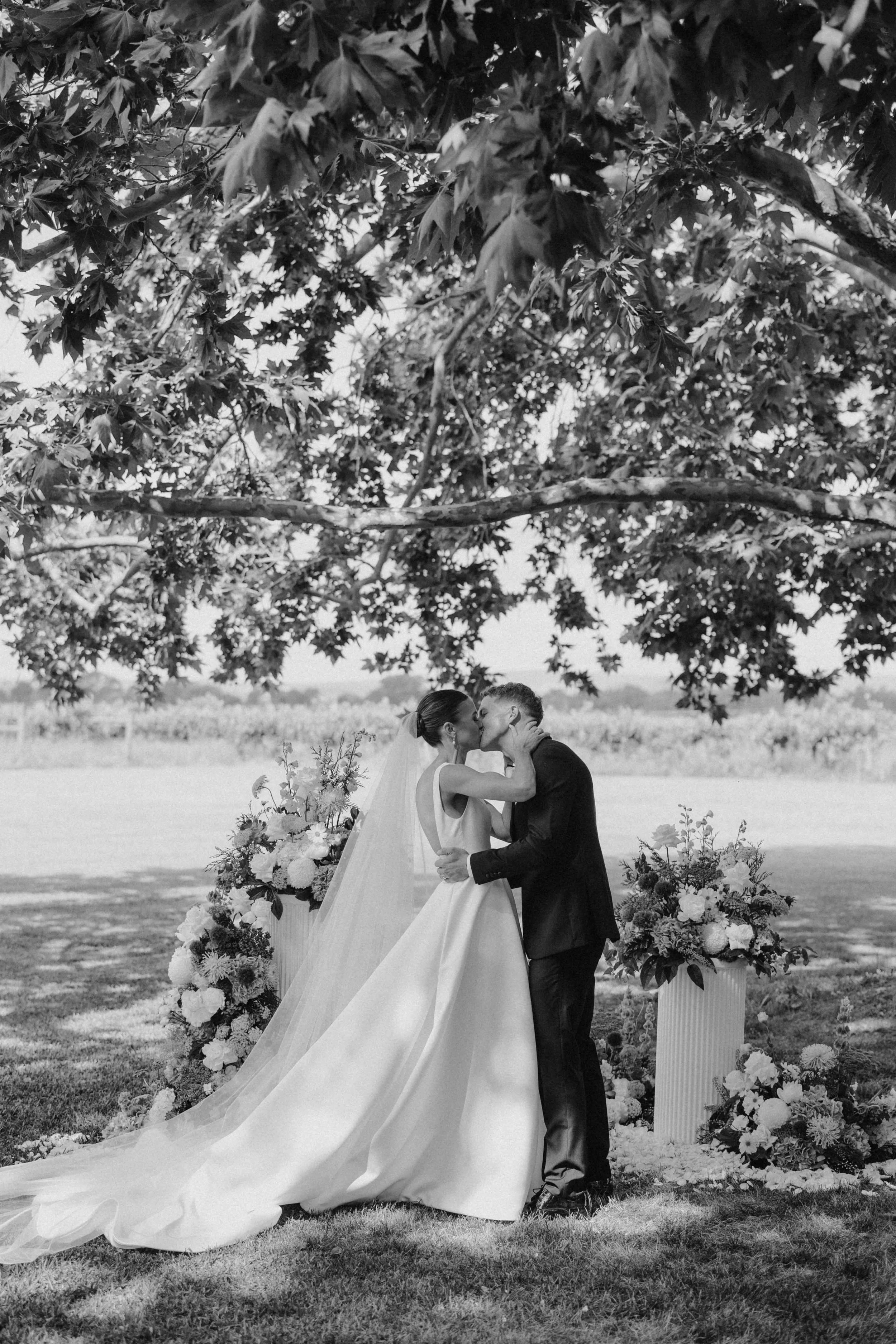 A black and white photograph of a bride and groom sharing a kiss outdoors beneath a tree, with floral arrangements on either side of them.