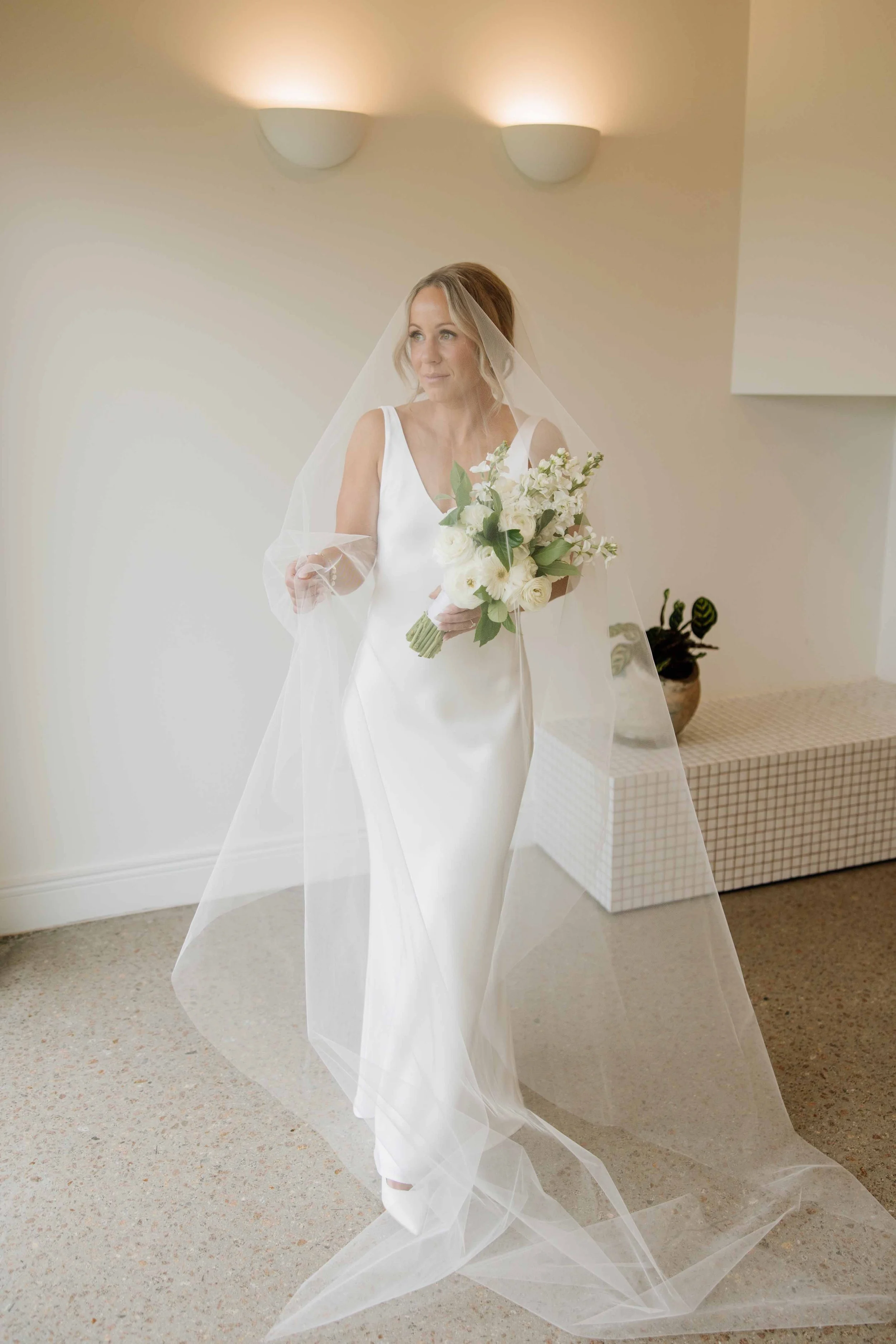 A bride standing indoors wearing a white wedding dress, holding a bouquet of white flowers, and looking to the left.
