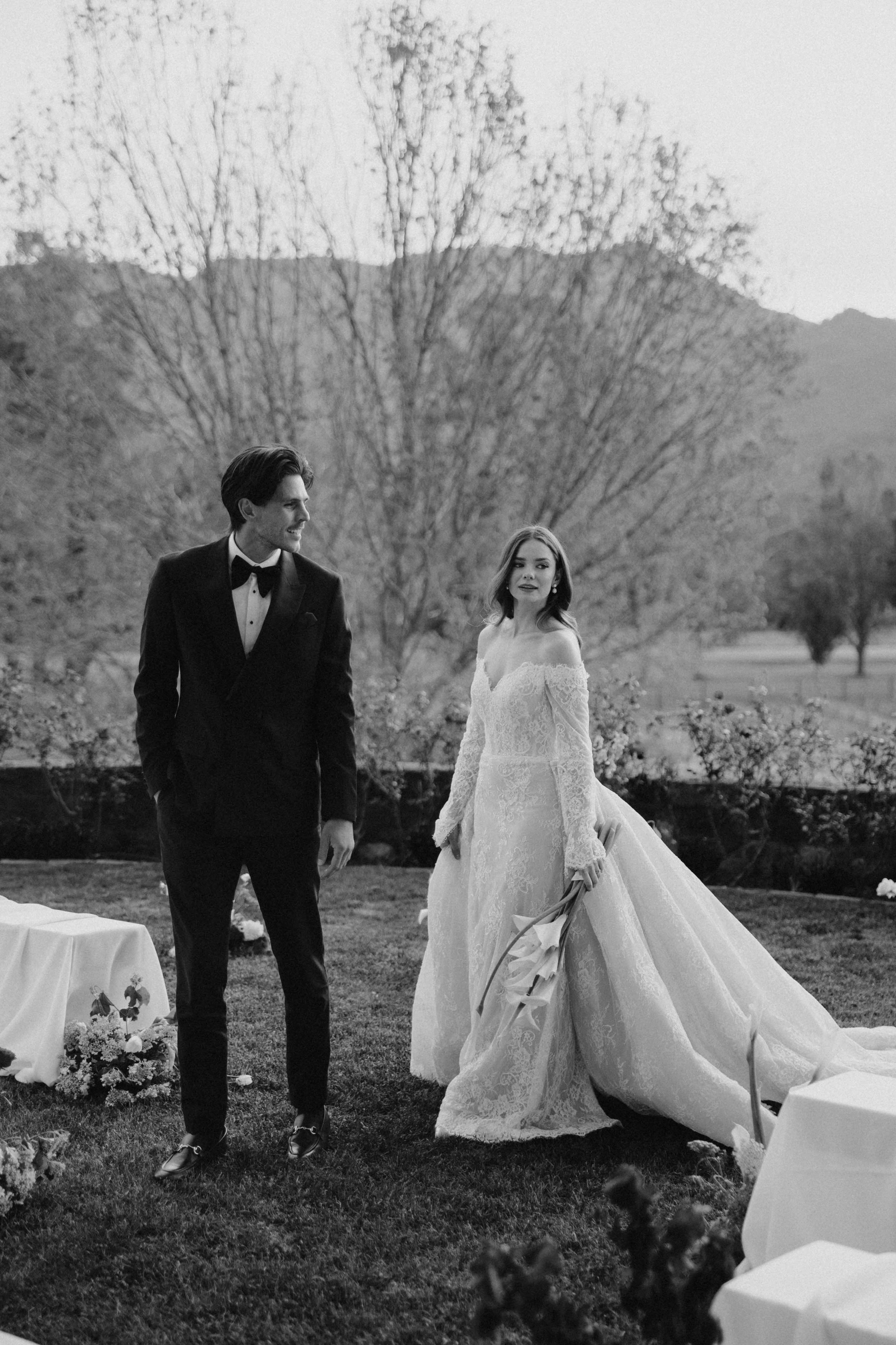 Black and white photo of a bride and groom outdoors, with the groom in a tuxedo and the bride holding a bouquet, surrounded by trees and tents.