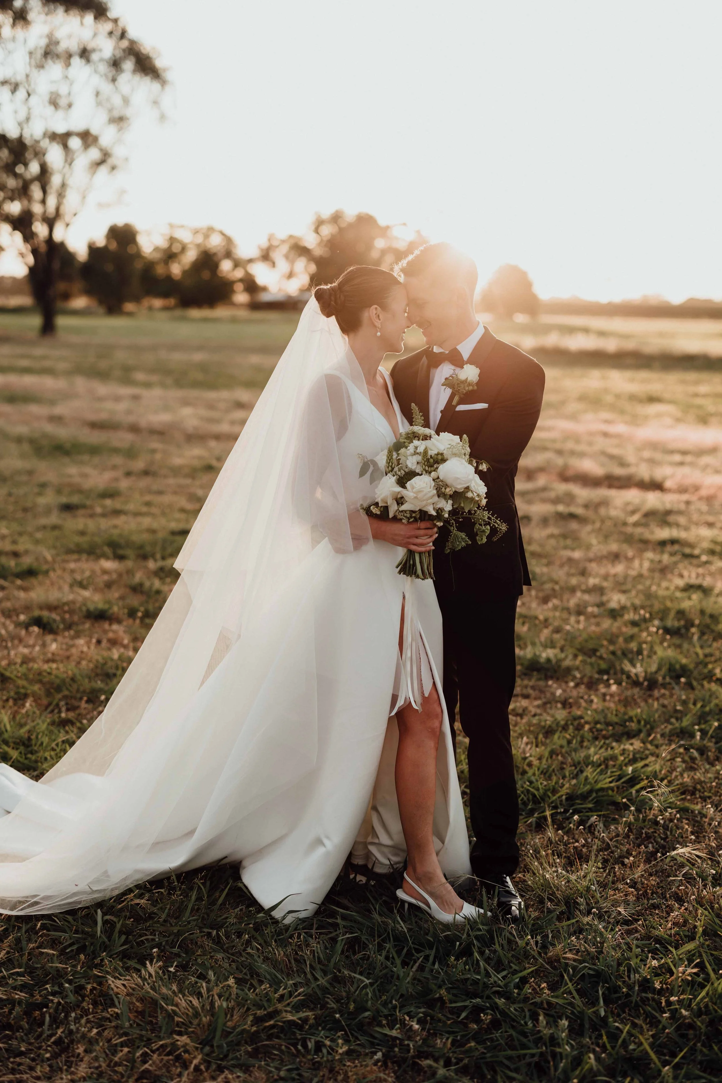 A bride and groom share a romantic moment outdoors during sunset, standing on grassy terrain with trees in the background. The bride wears a white wedding gown, veil, and high heels, holding a bouquet of white flowers. The groom is dressed in a black tuxedo with a bowtie, with their foreheads touching.
