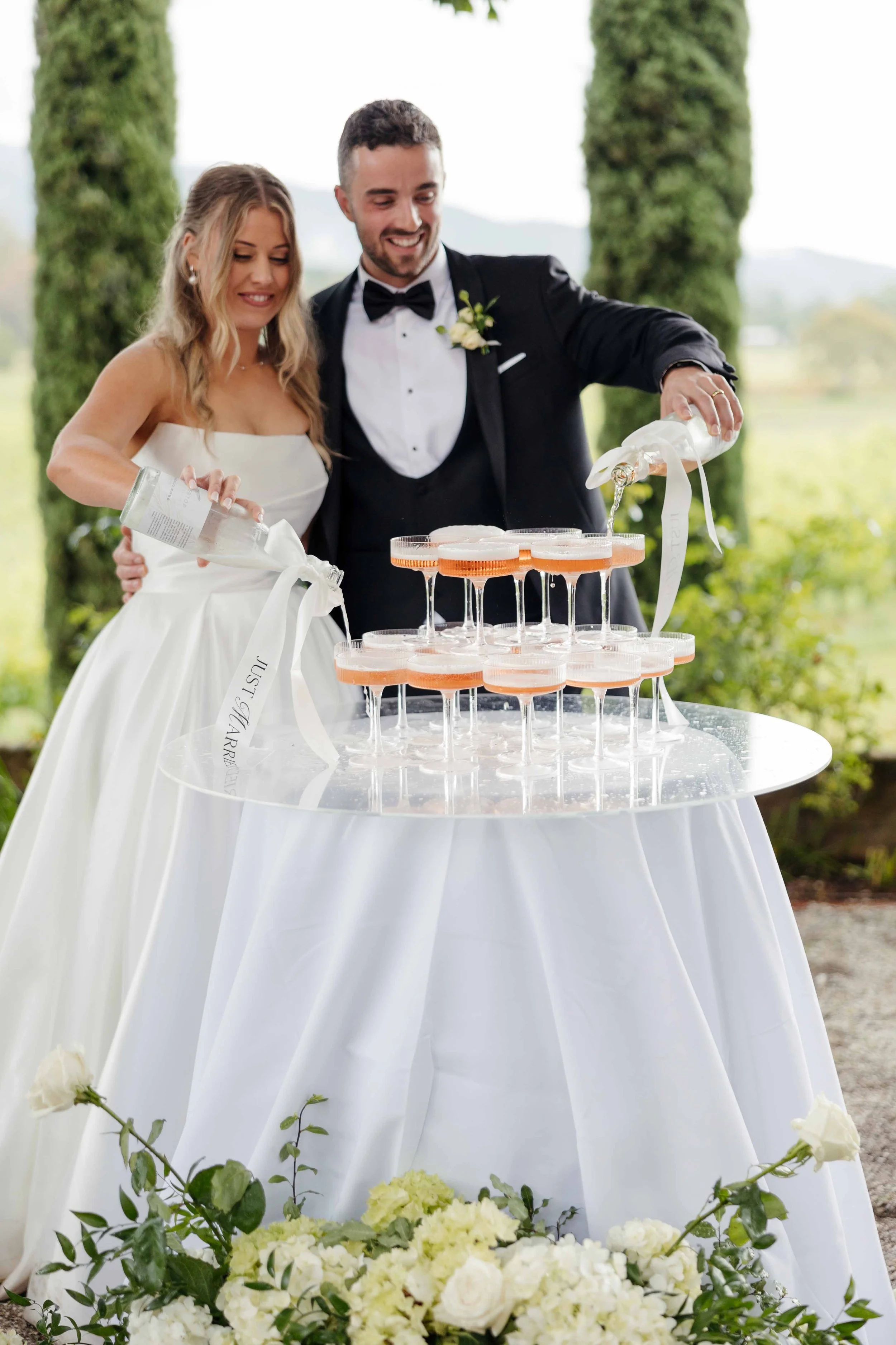 Bride and groom at a wedding pouring champagne into glasses on a tiered stand, outdoors with greenery.
