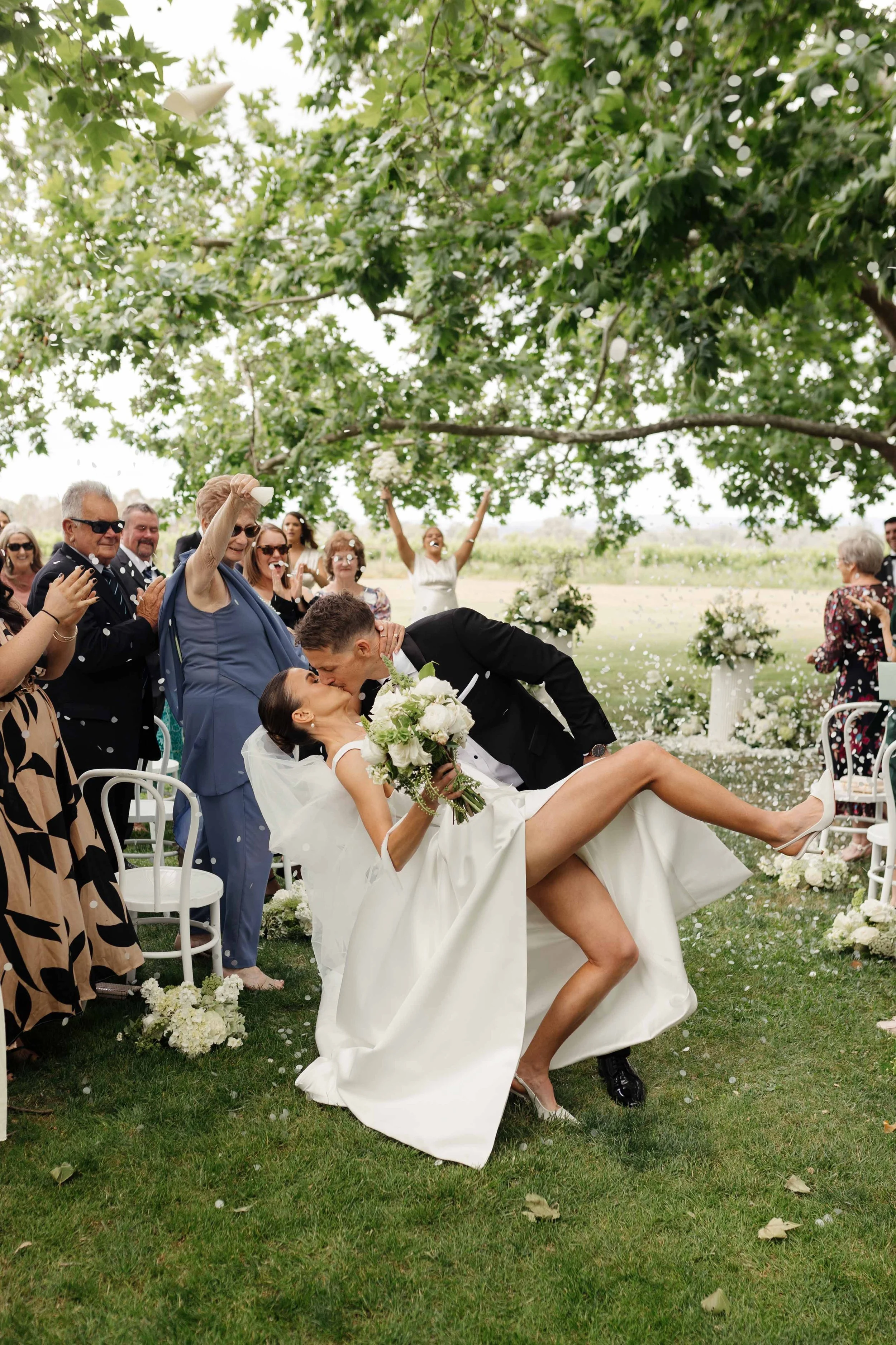 A wedding celebration outdoors with a bride and groom, where the groom is kissing the bride who is holding a bouquet, while guests cheer and throw confetti beneath a large tree.