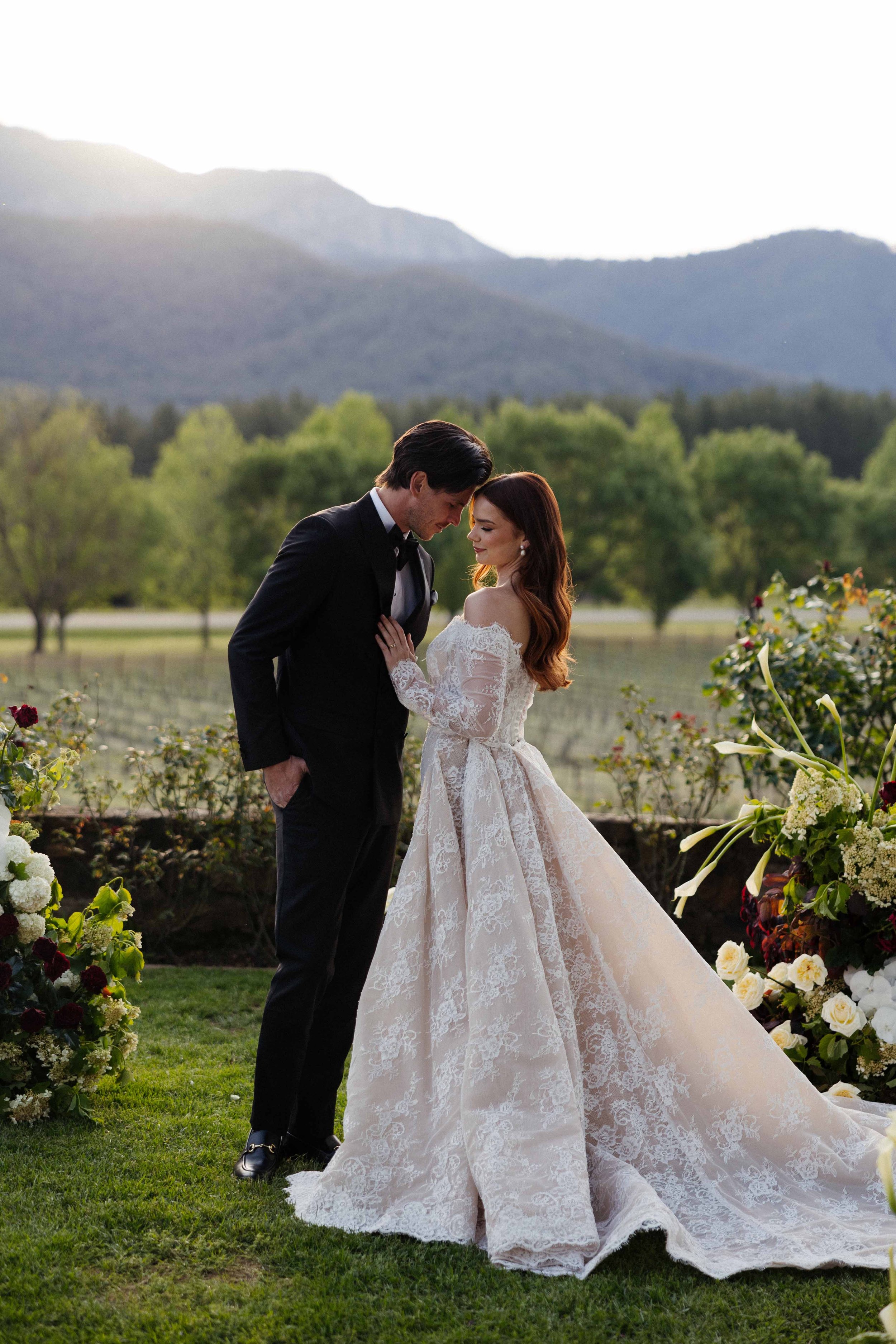 A bride and groom stand closely together outdoors, surrounded by floral arrangements, with mountains and trees in the background during sunset.