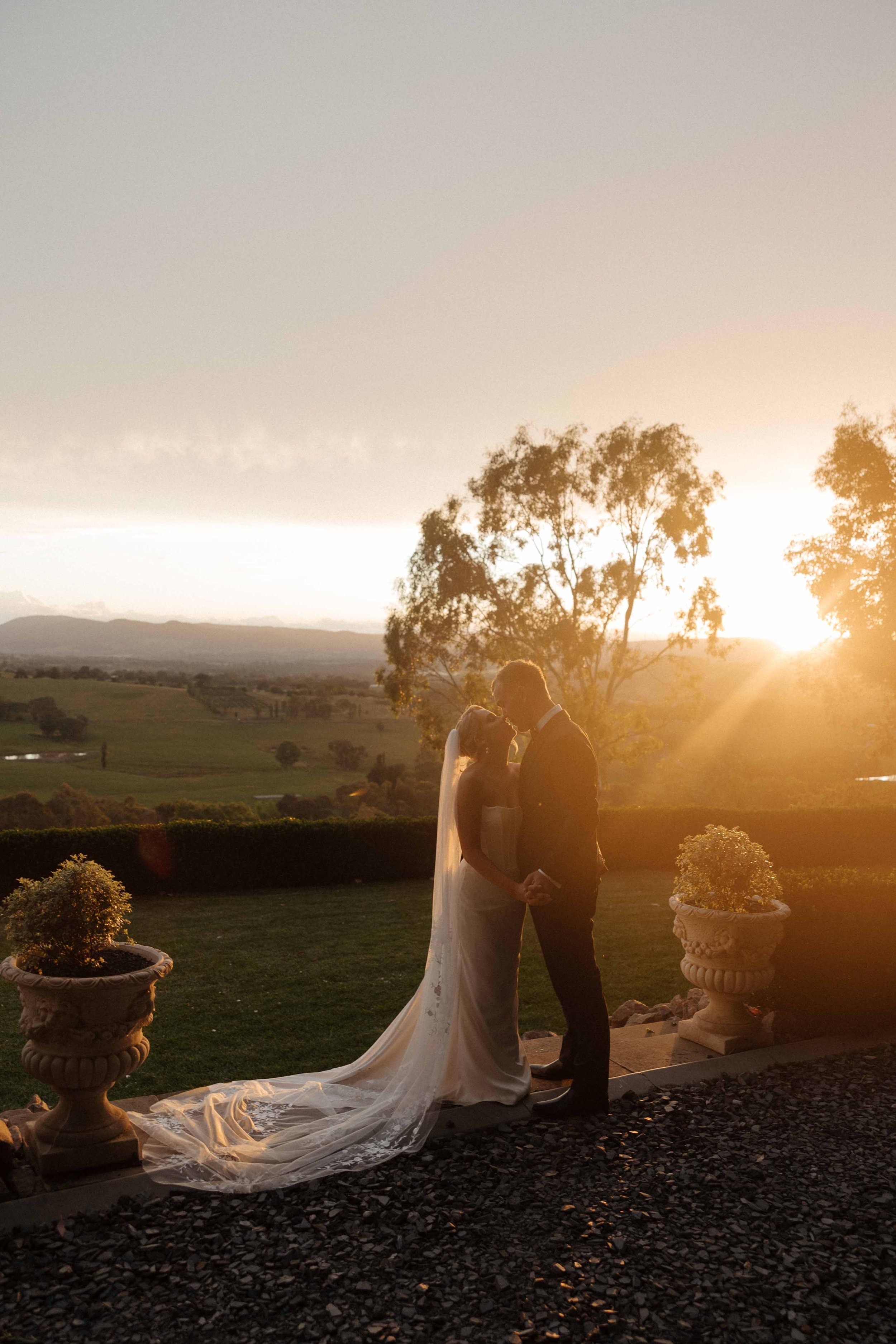 A bride and groom share a kiss during their wedding at sunset, with the sun setting behind trees and an open landscape in the background.