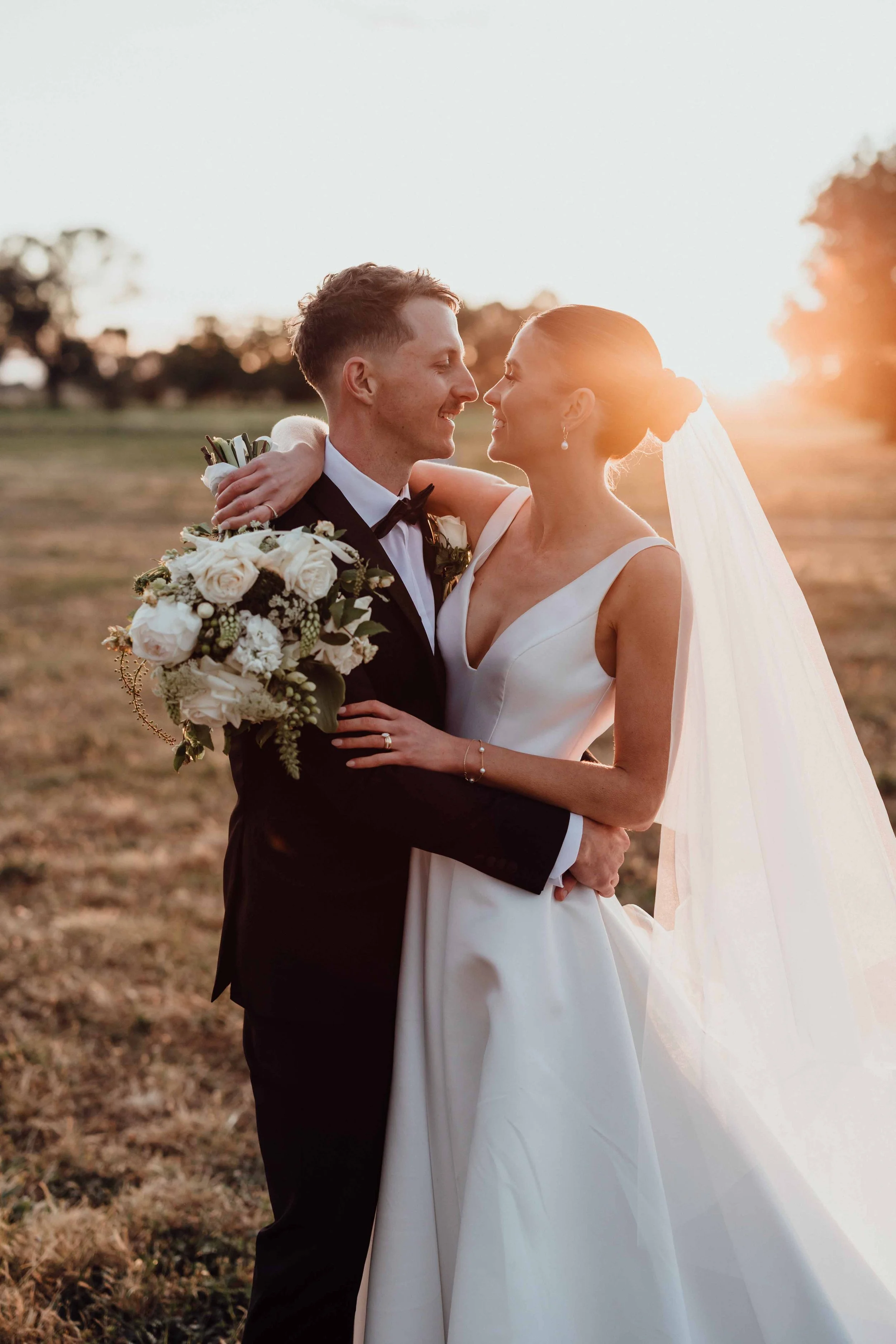Bride and groom embracing outdoors during sunset, the bride in a white wedding gown with a long veil, and the groom in a black tuxedo, holding a bouquet of white flowers.