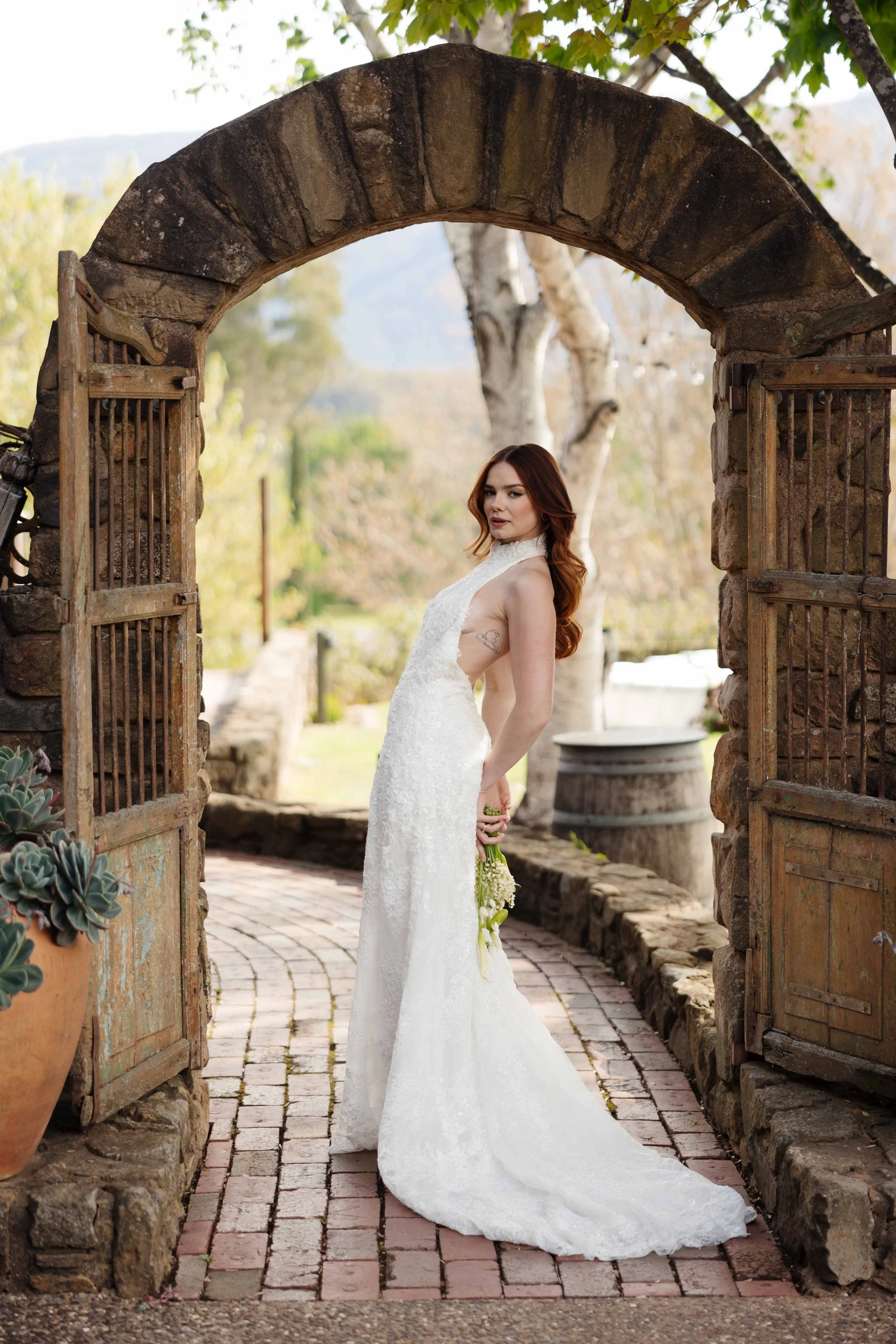 A bride in a white lace wedding dress holding a small bouquet of white flowers, standing under a rustic stone and wood archway outside.