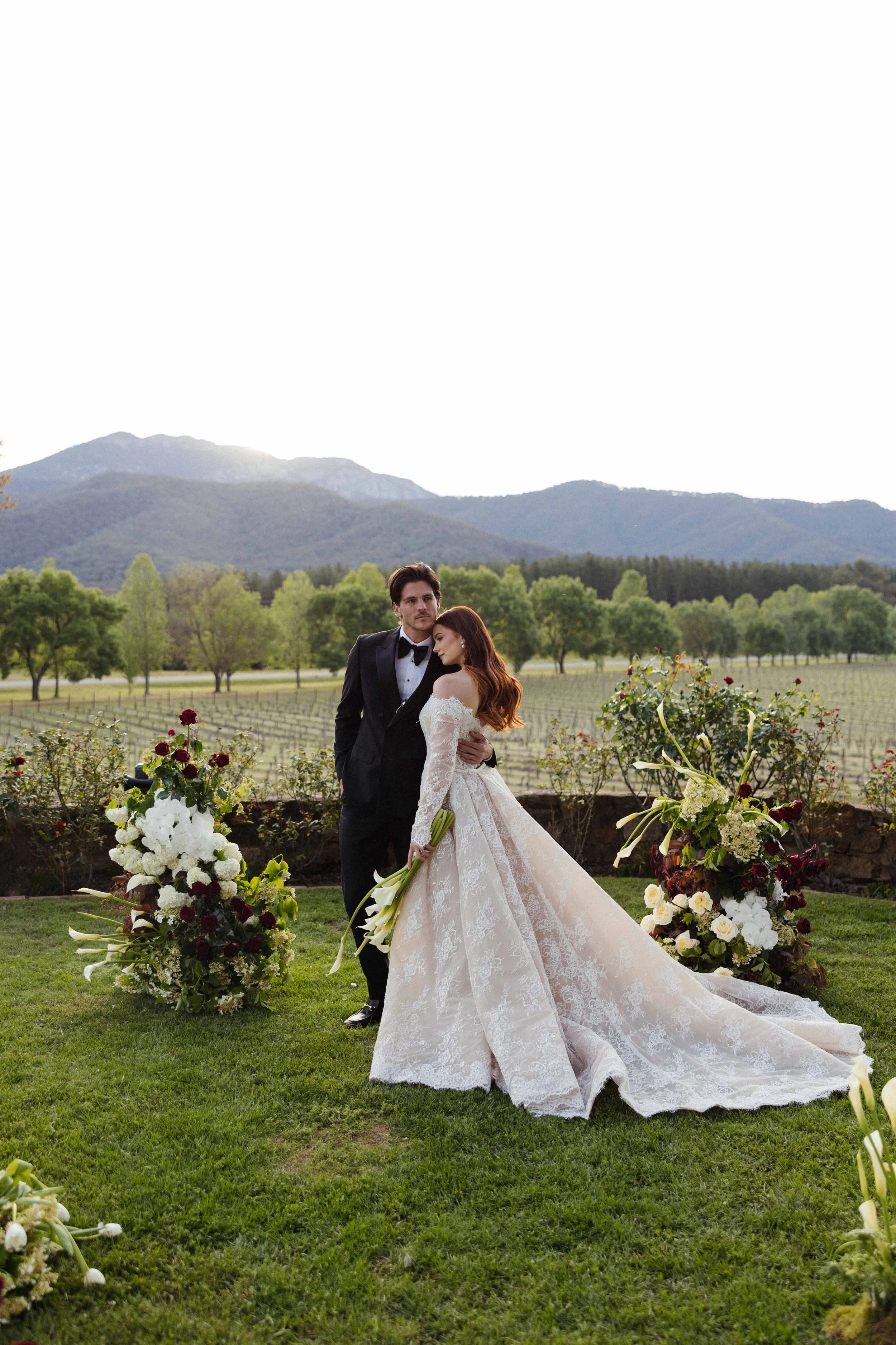 A bride and groom standing together outdoors during their wedding, with mountains and trees in the background. The bride is holding a bouquet of white calla lilies and wearing a lace wedding gown. The groom is dressed in a black tuxedo. Floral arrangements are on either side of them.