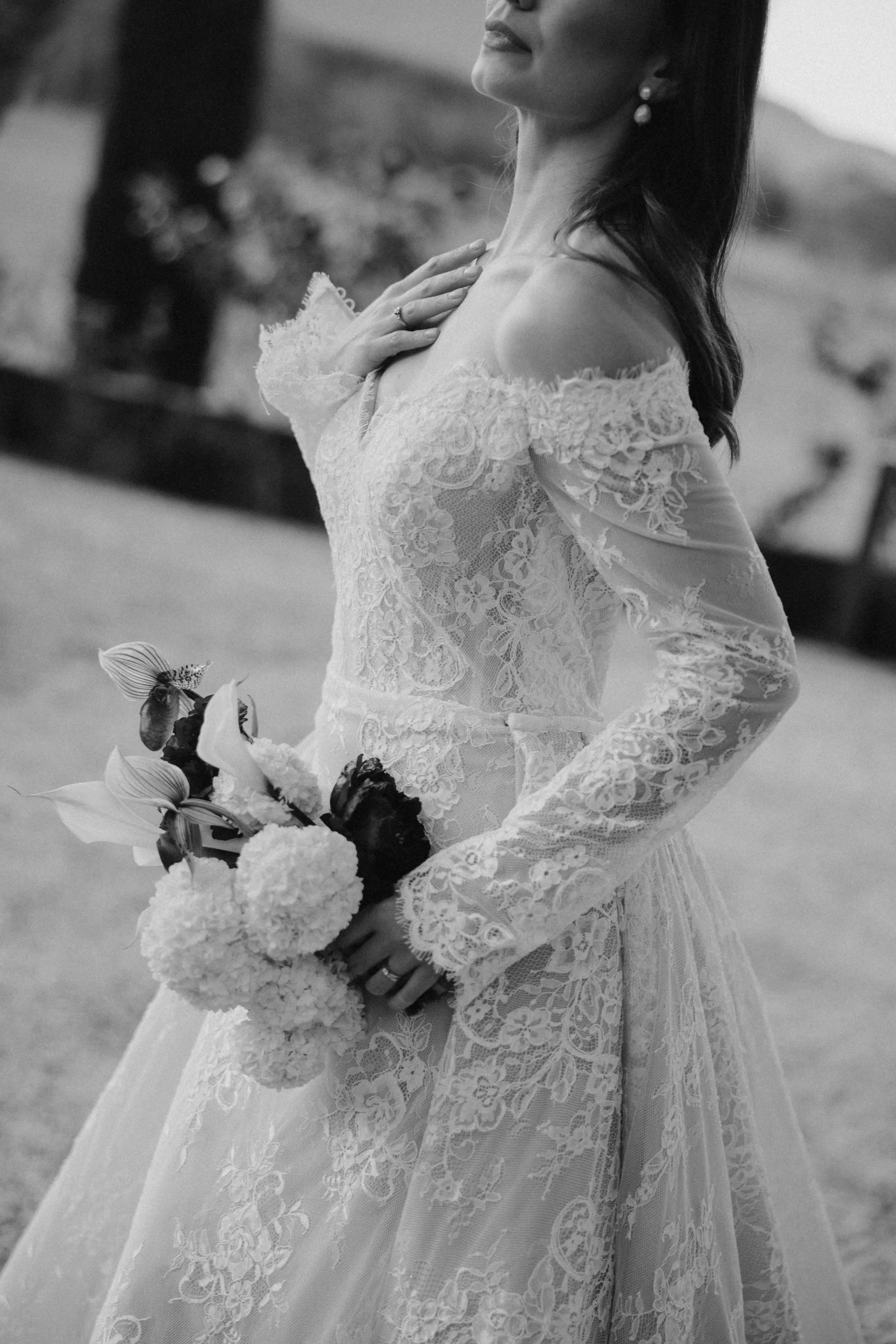 A bride in a lace wedding dress holding a bouquet of flowers, standing outdoors.
