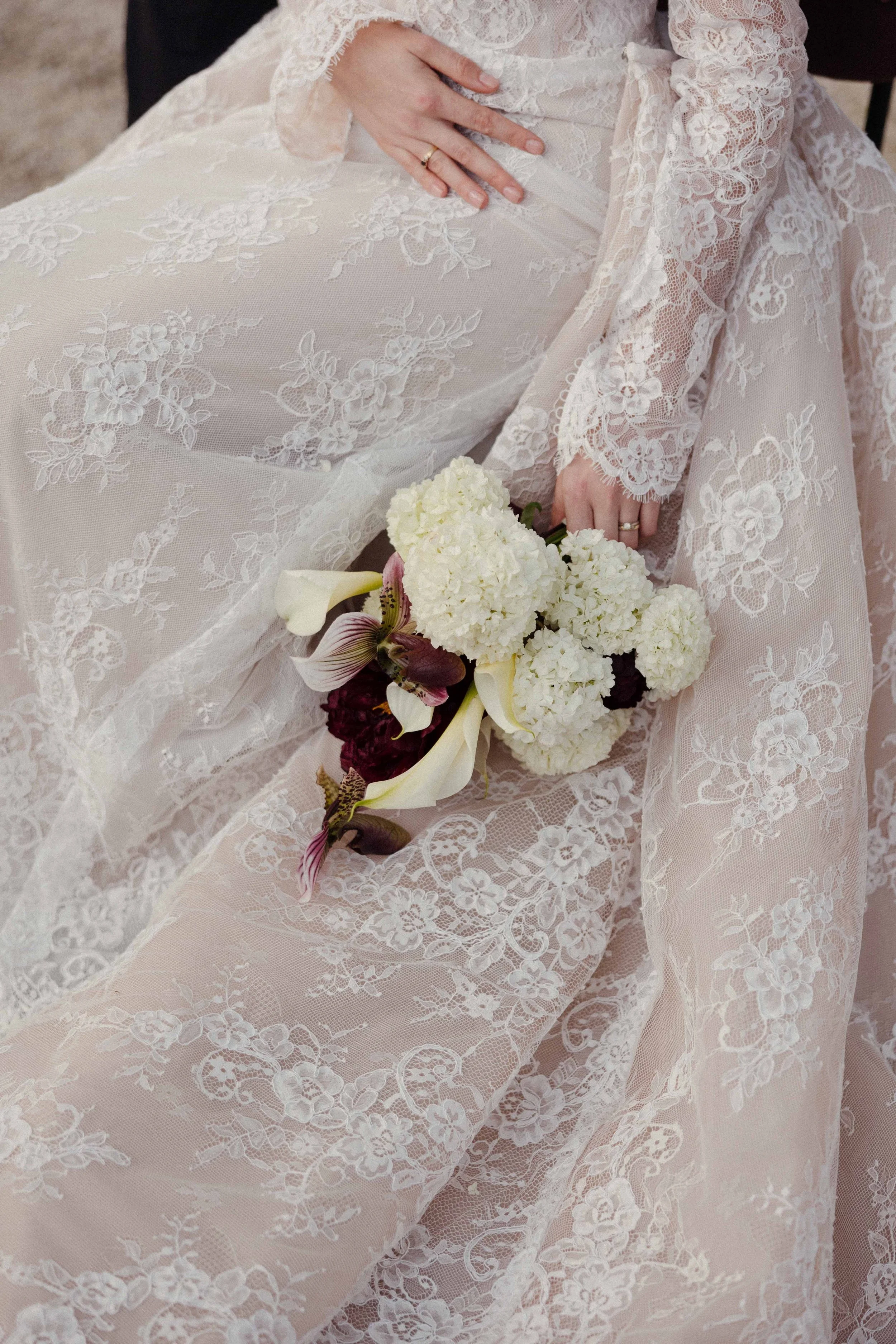 A person wearing a long, white lace wedding dress holding a bouquet of white and dark red flowers.
