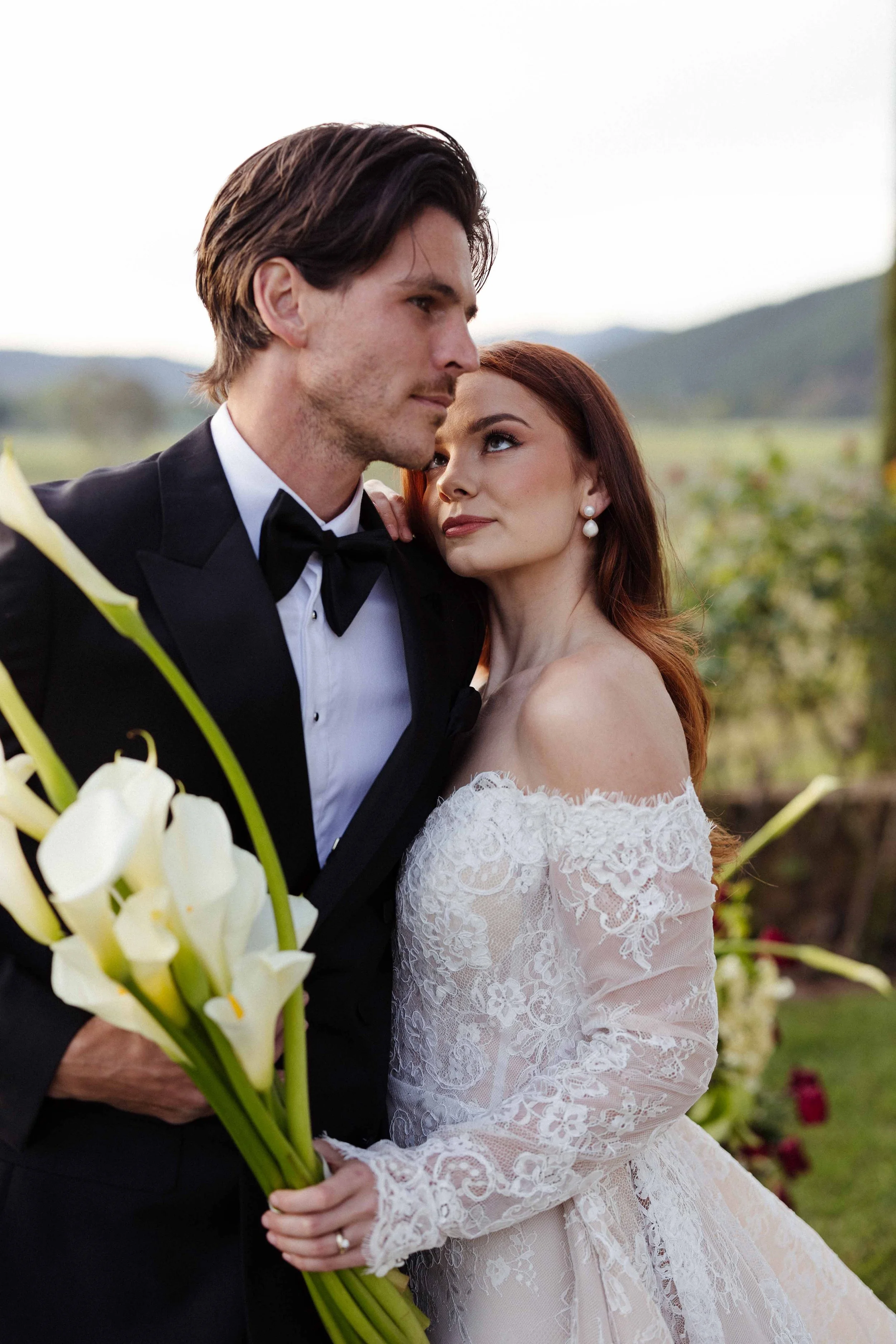 A bride and groom in wedding attire embrace outdoors, with a bride holding a bouquet of white calla lilies, surrounded by greenery and mountains in the background.