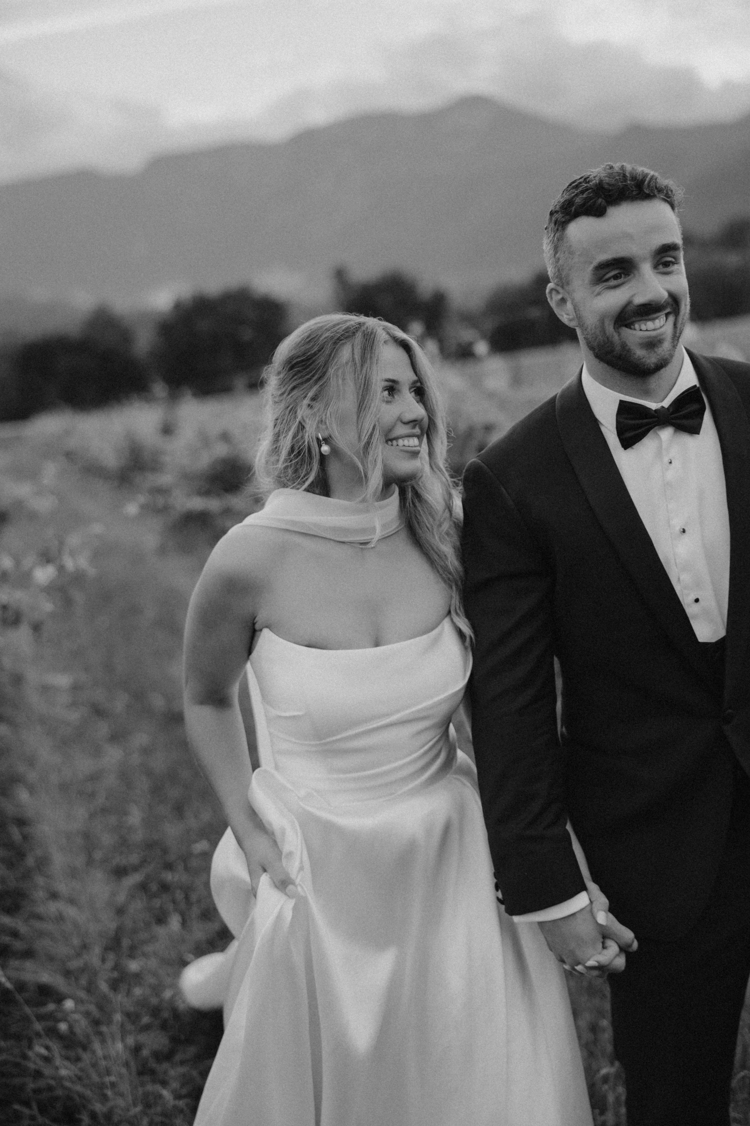 A bride and groom holding hands outdoors, smiling, with mountains and trees in the background, black and white photo.