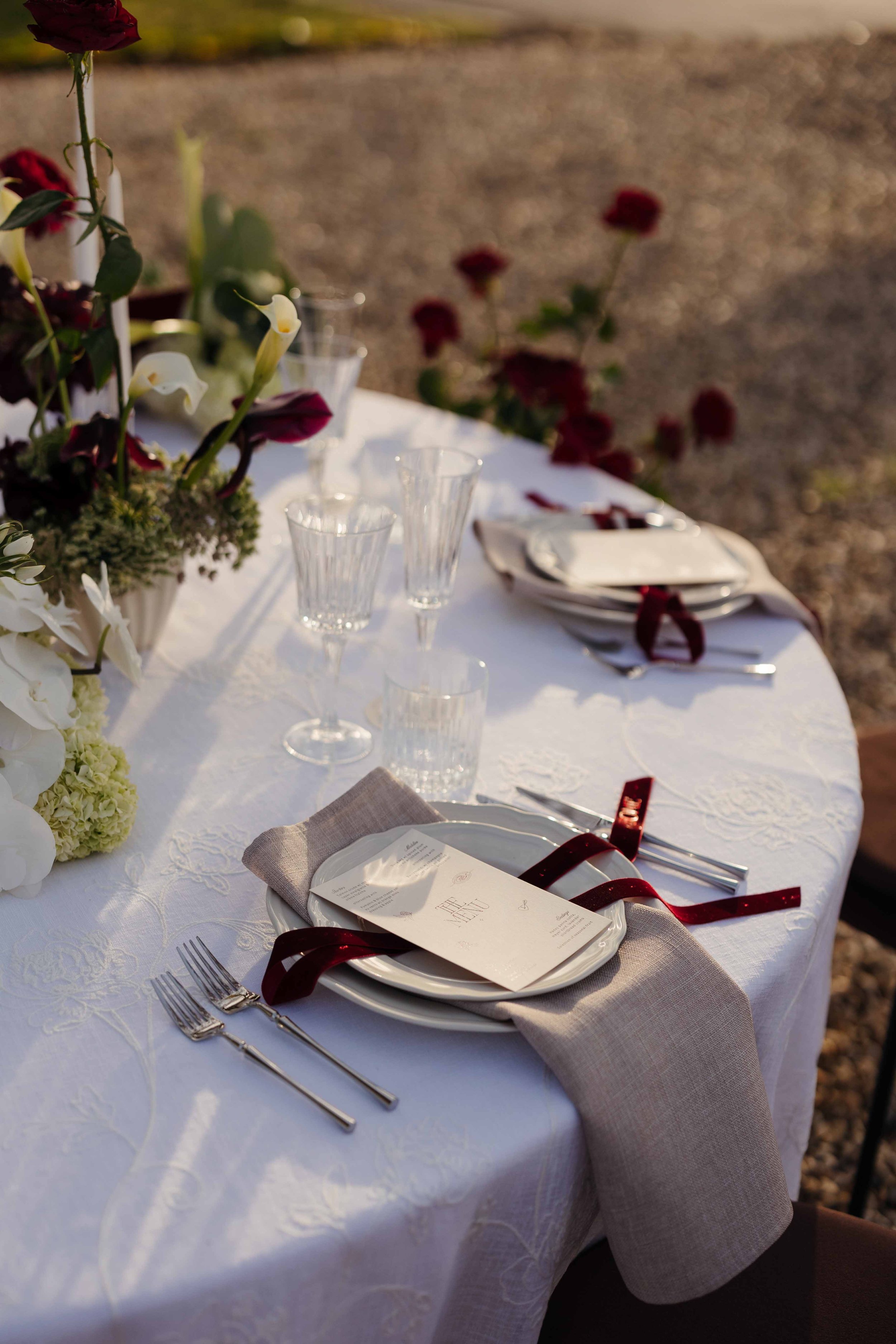 Elegant outdoor table set for a special occasion with white tablecloth, floral centerpiece, wine glasses, plates, silverware, and a menu, decorated with red ribbons in the late afternoon.