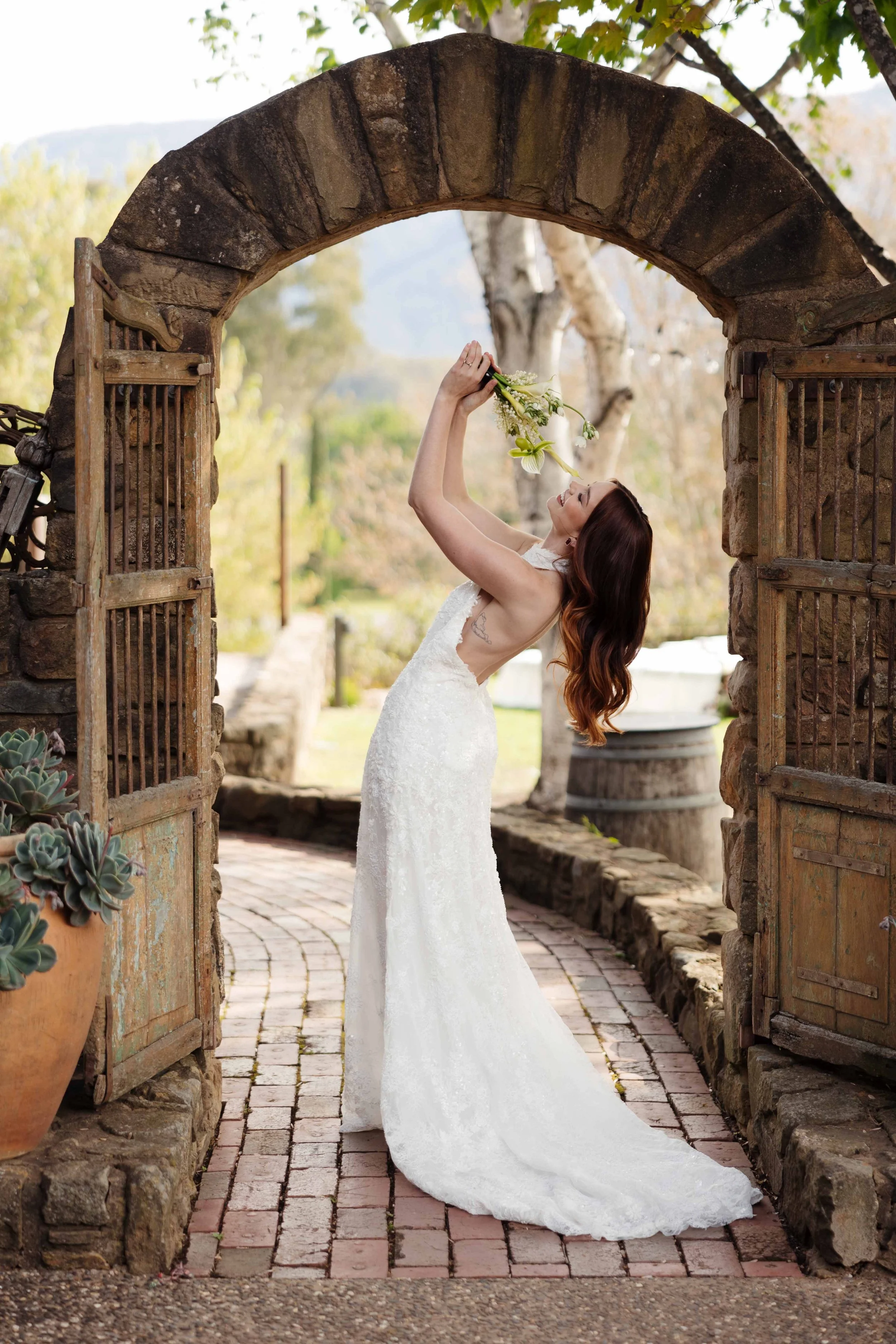 A woman in a white wedding dress is holding a bouquet of flowers above her head and leaning back in an outdoor setting with stone and wooden gate archway, greenery, and trees in the background.