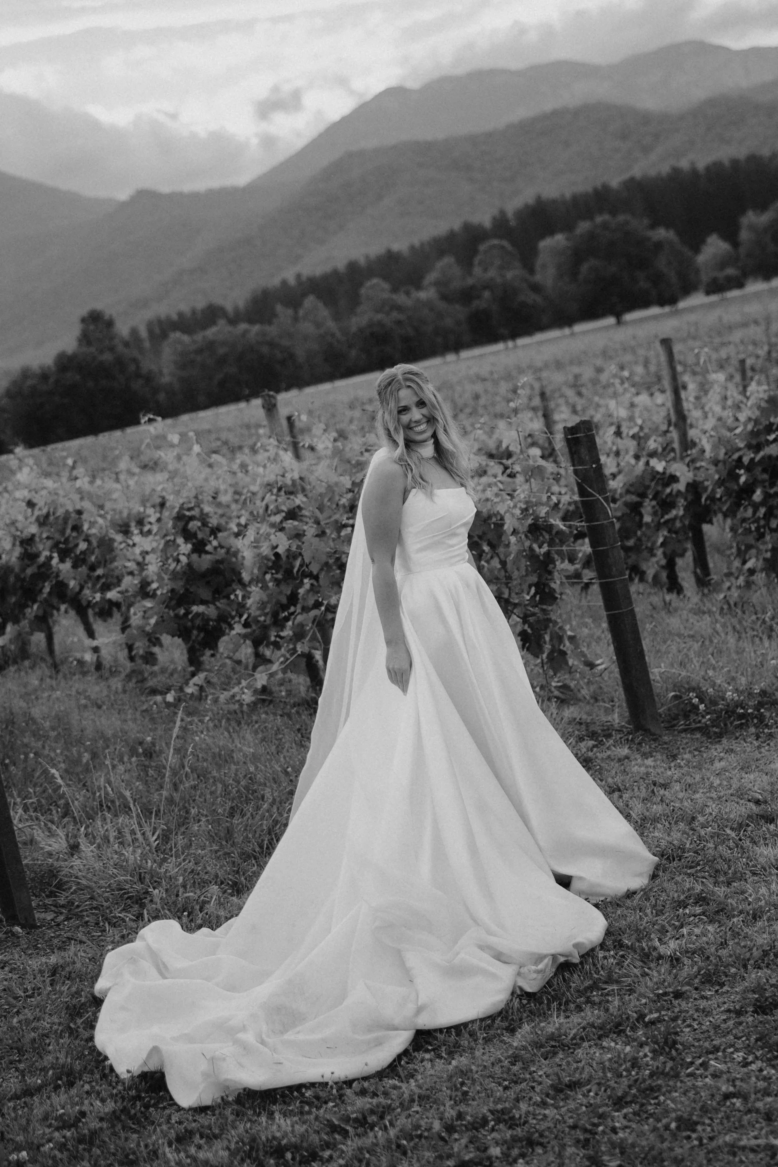 A smiling bride in a strapless wedding gown with a long train standing in a vineyard with mountains in the background.