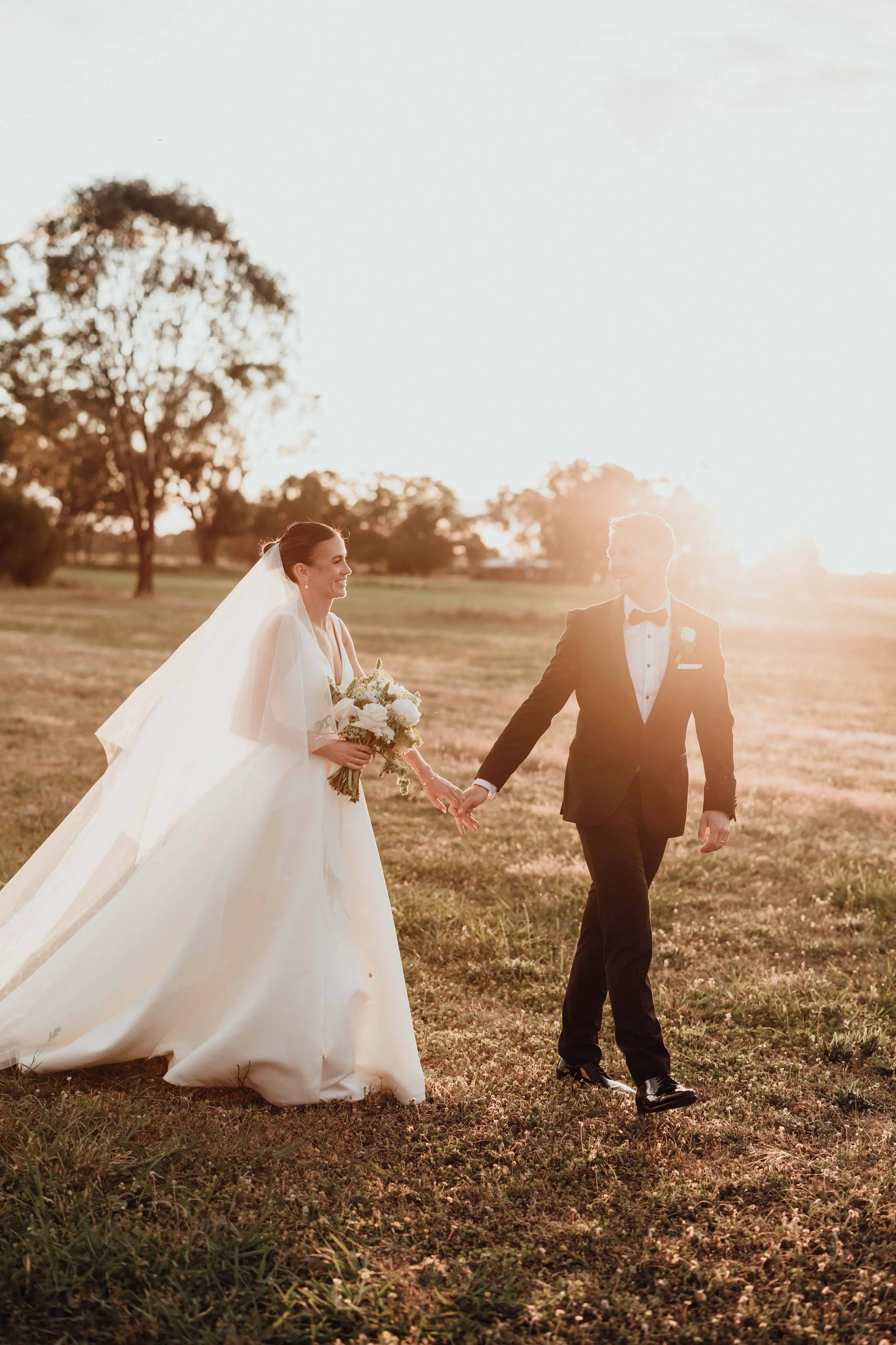A bride and groom holding hands and smiling at each other outdoors during sunset, with the bride wearing a white wedding gown and holding a bouquet, and the groom in a black tuxedo.
