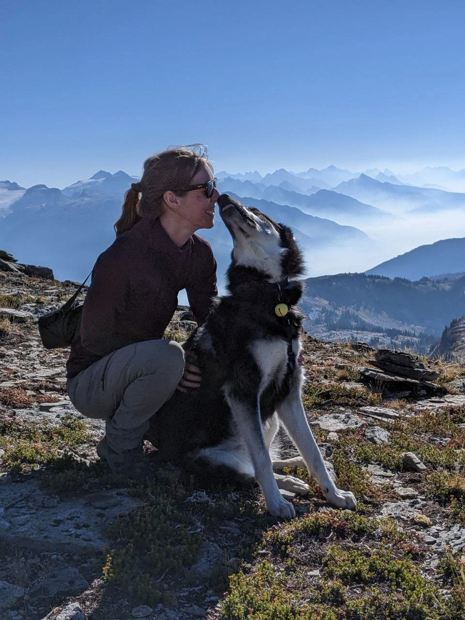 A woman and a Siberian Husky dog on a mountain trail with a backdrop of mountains and a clear blue sky. The woman is squatting and leaning towards the dog, which appears to be licking her face.
