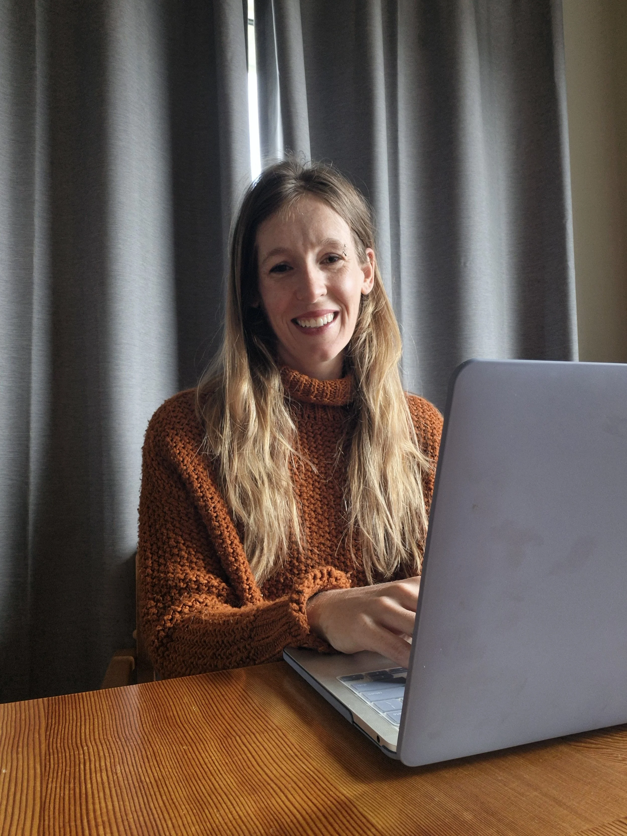 A woman with long blonde hair, smiling, sitting at a wooden table using a silver laptop, with gray curtains in the background.
