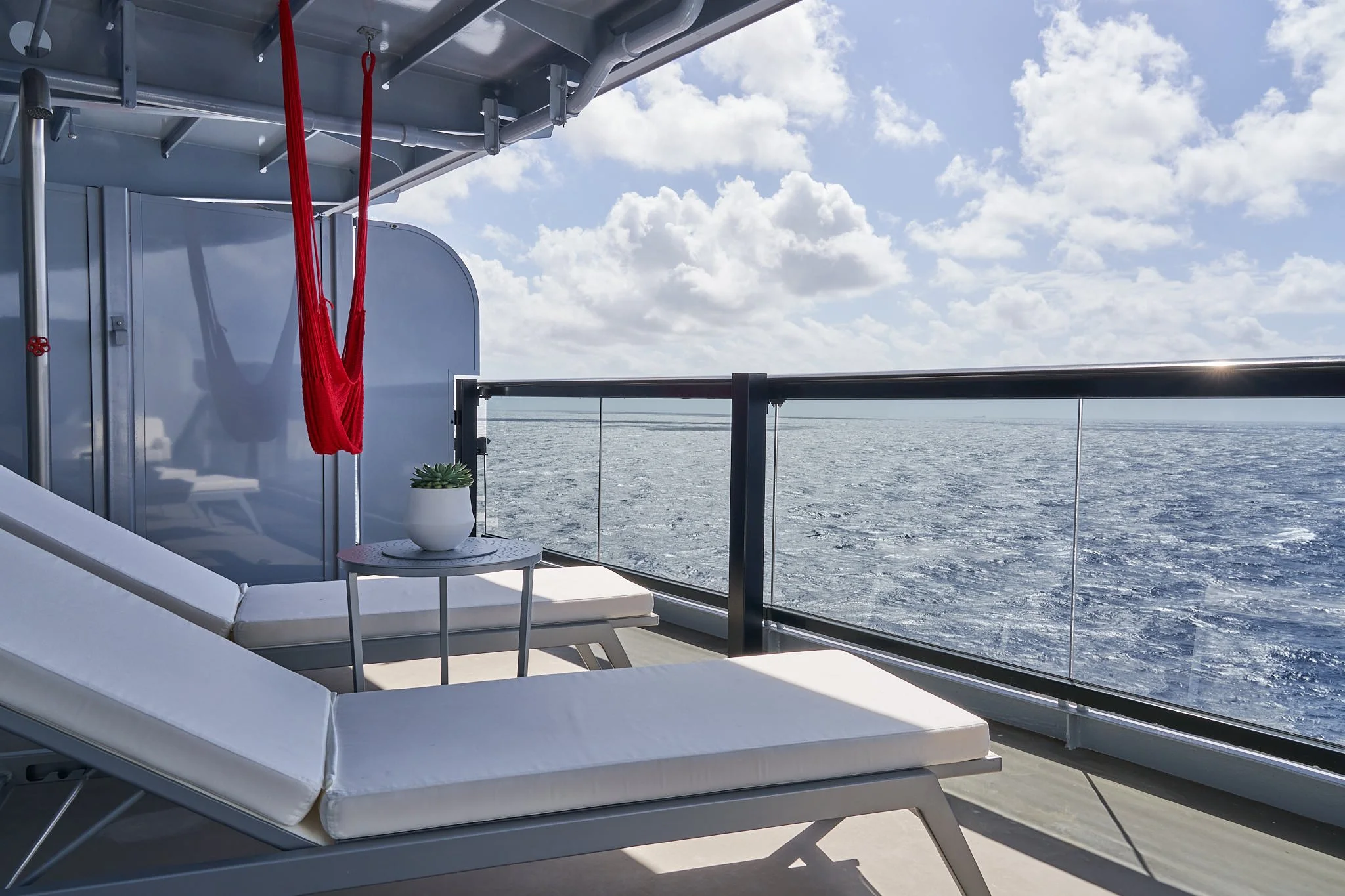 View of a cruise ship balcony with two white lounge chairs, a small table with a potted succulent plant, a red hammock, and a glass railing overlooking the ocean under a partly cloudy sky.