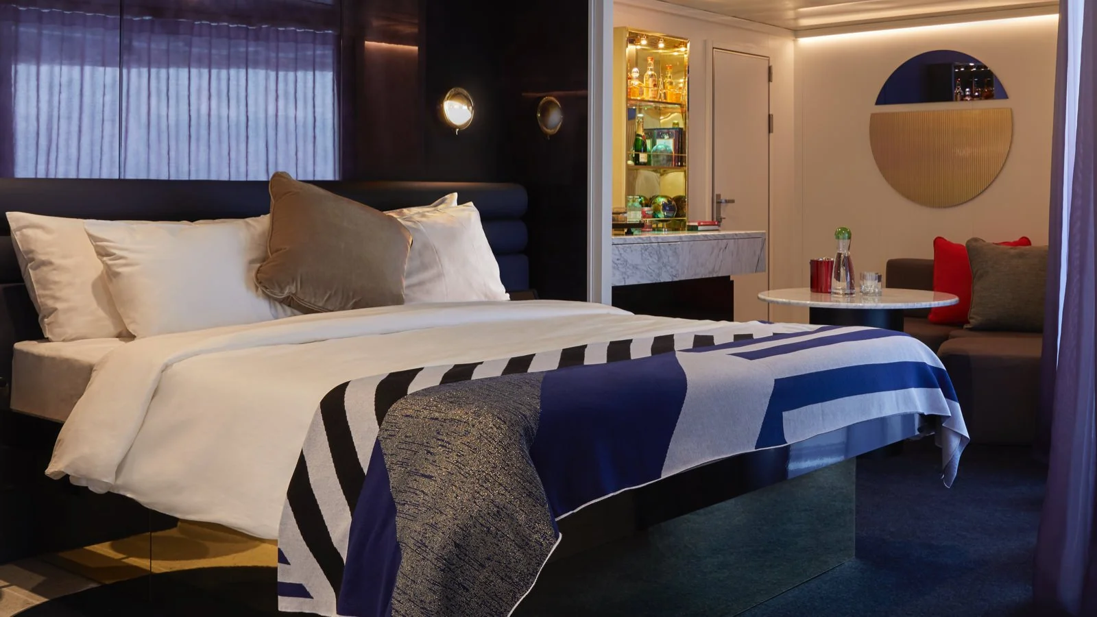 A stylish hotel room with a large bed, pillows, and a patterned blanket. There is a small round table with a water bottle and cups, a sitting area with cushions, and a minibar or liquor cabinet in the corner. The room has modern decor and soft lighting.