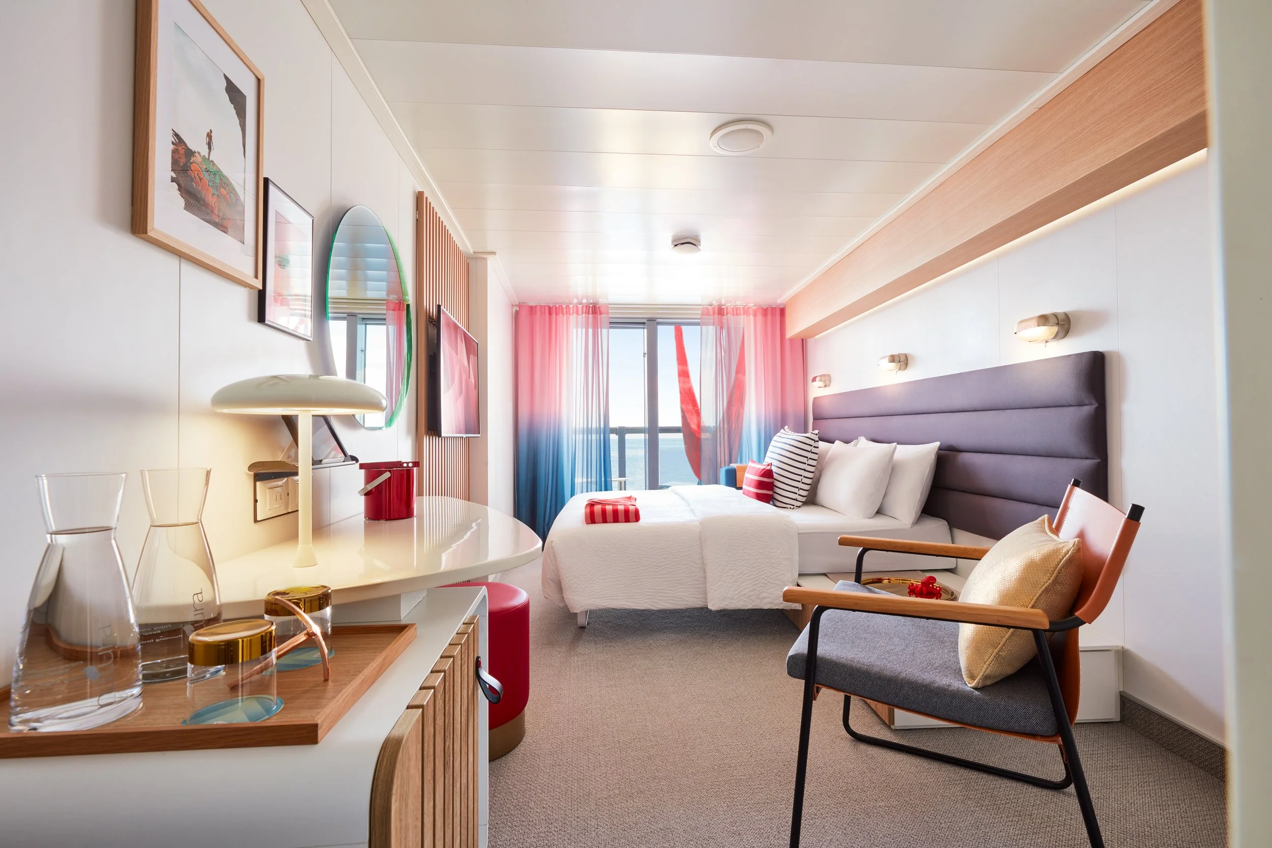 A cozy hotel room with a modern decor featuring a bed with white linens and striped pillows, a sitting chair with a cushion, a wall-mounted television, and a window with pink and blue curtains overlooking the ocean.