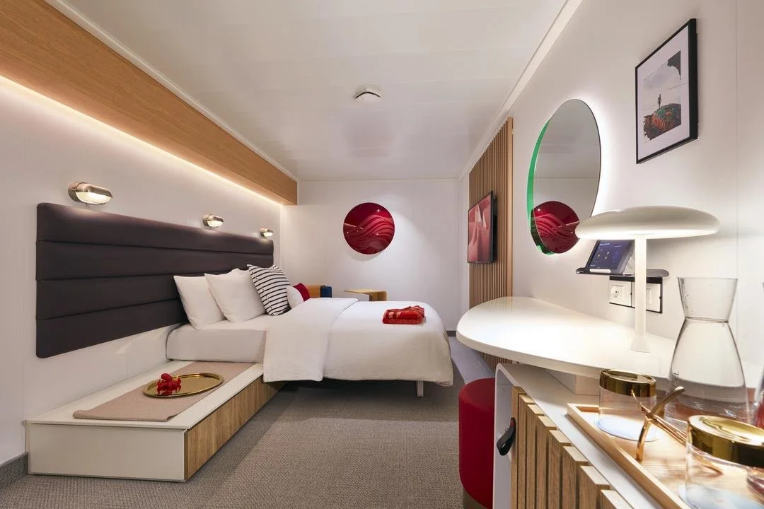 Modern hotel room with a large bed, white bedding, black and white pillows, wall-mounted lights, a red circular art piece, a large mirror, a flat-screen TV, a white desk with a modern lamp, and decorative items.