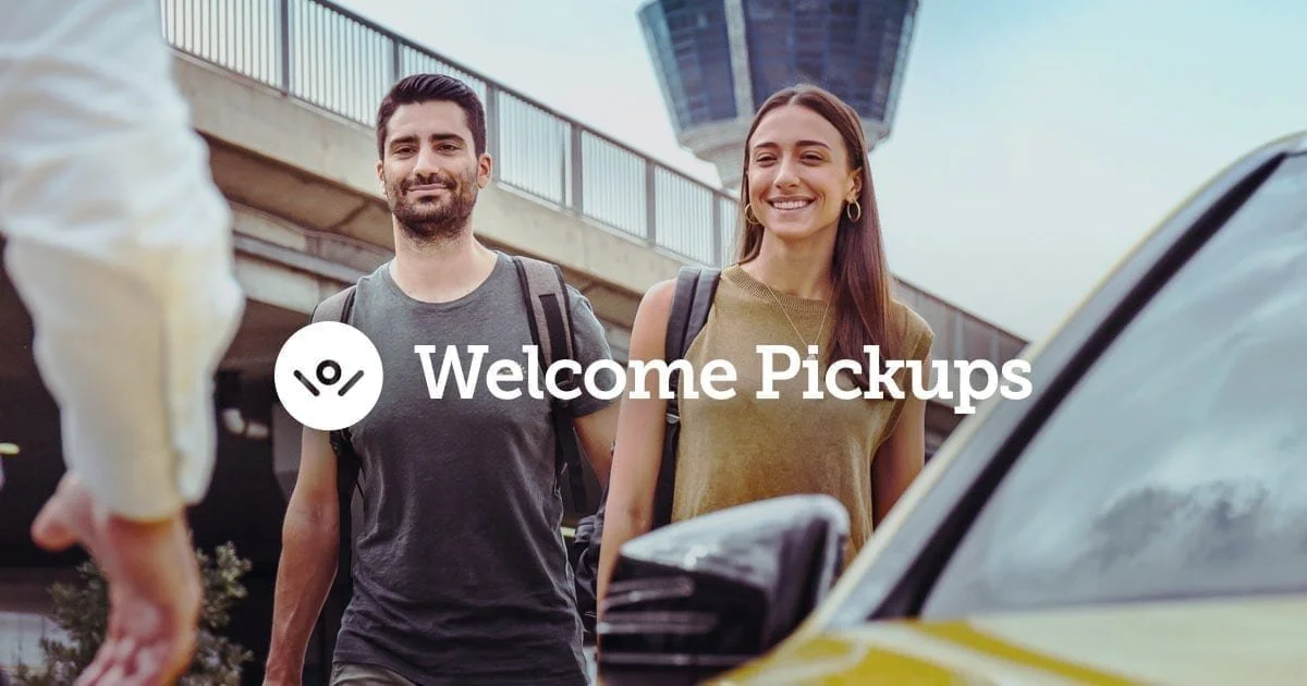 A man and woman with backpacks arriving at a ride-sharing pickup point in an urban area, smiling.