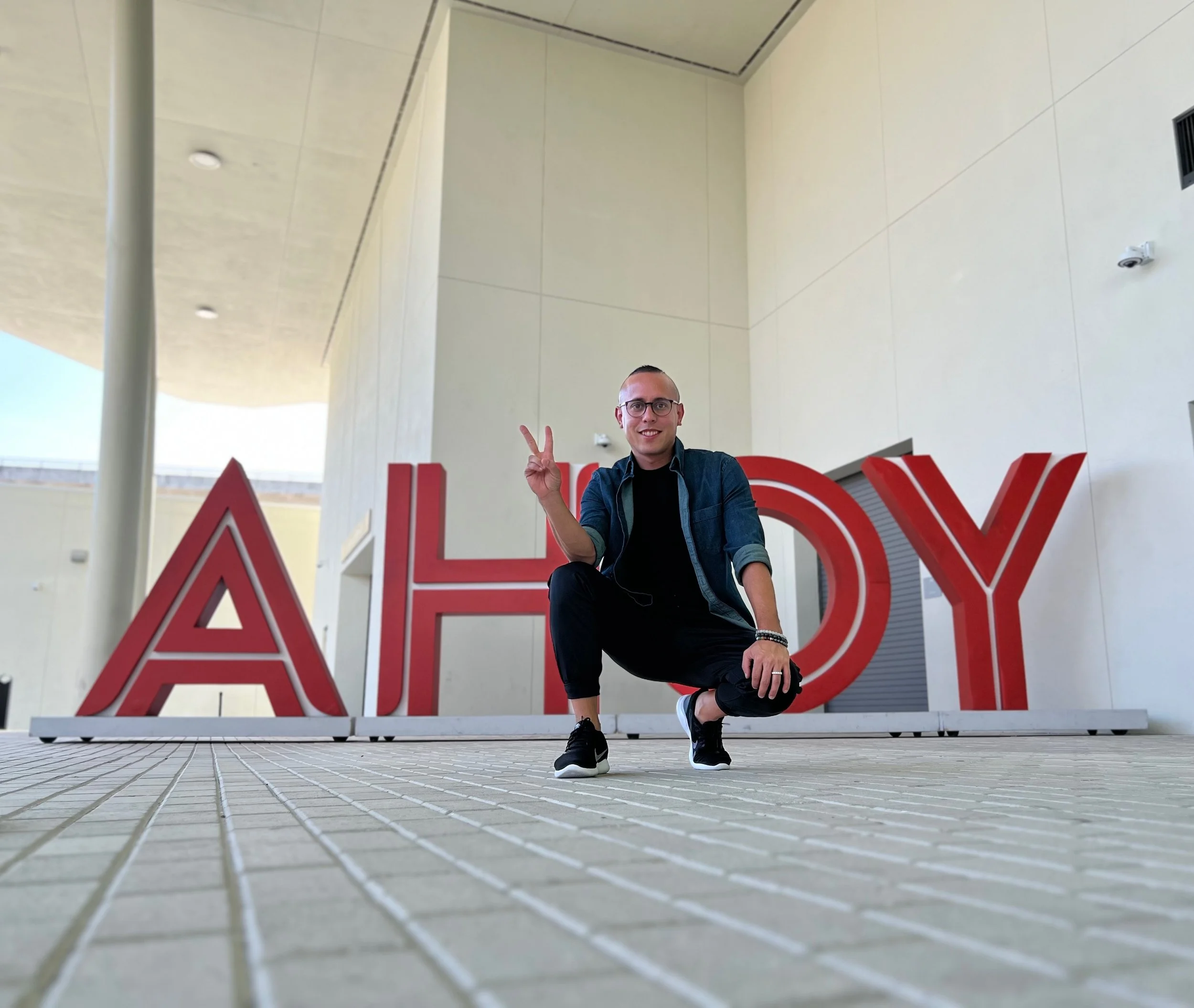 A young man crouching in front of large red letters spelling 'ALOHA' at an outdoor location with modern building architecture in the background.