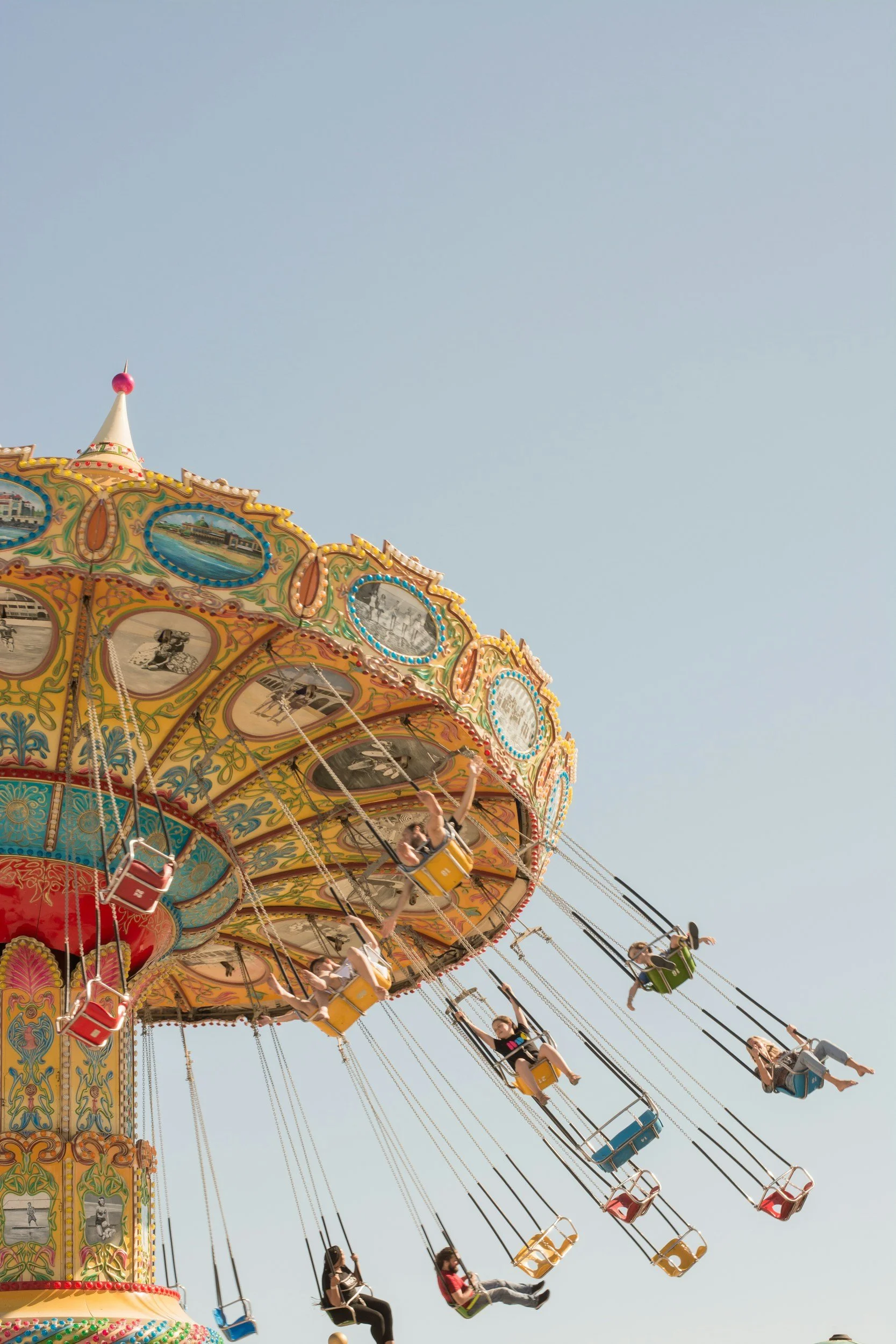 Children riding on a colorful carousel swing ride at an amusement park against a clear blue sky.