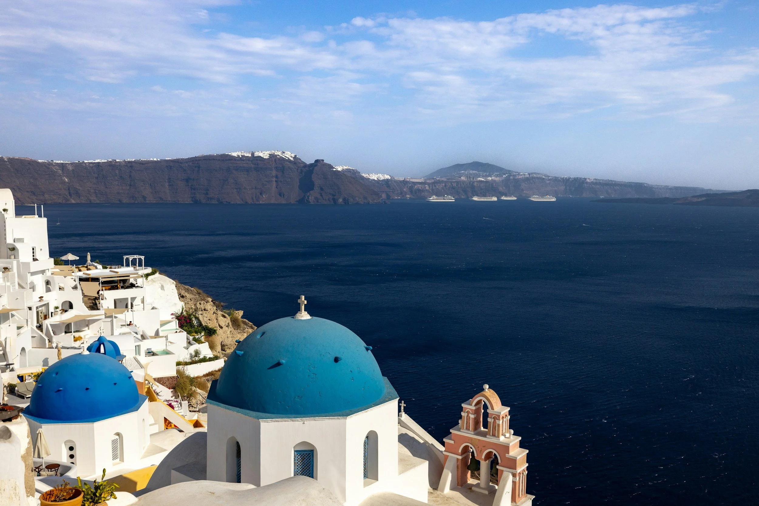 A view of the ocean with blue-domed churches and white buildings in Santorini, Greece, with cruise ships in the distance and mountains across the water.
