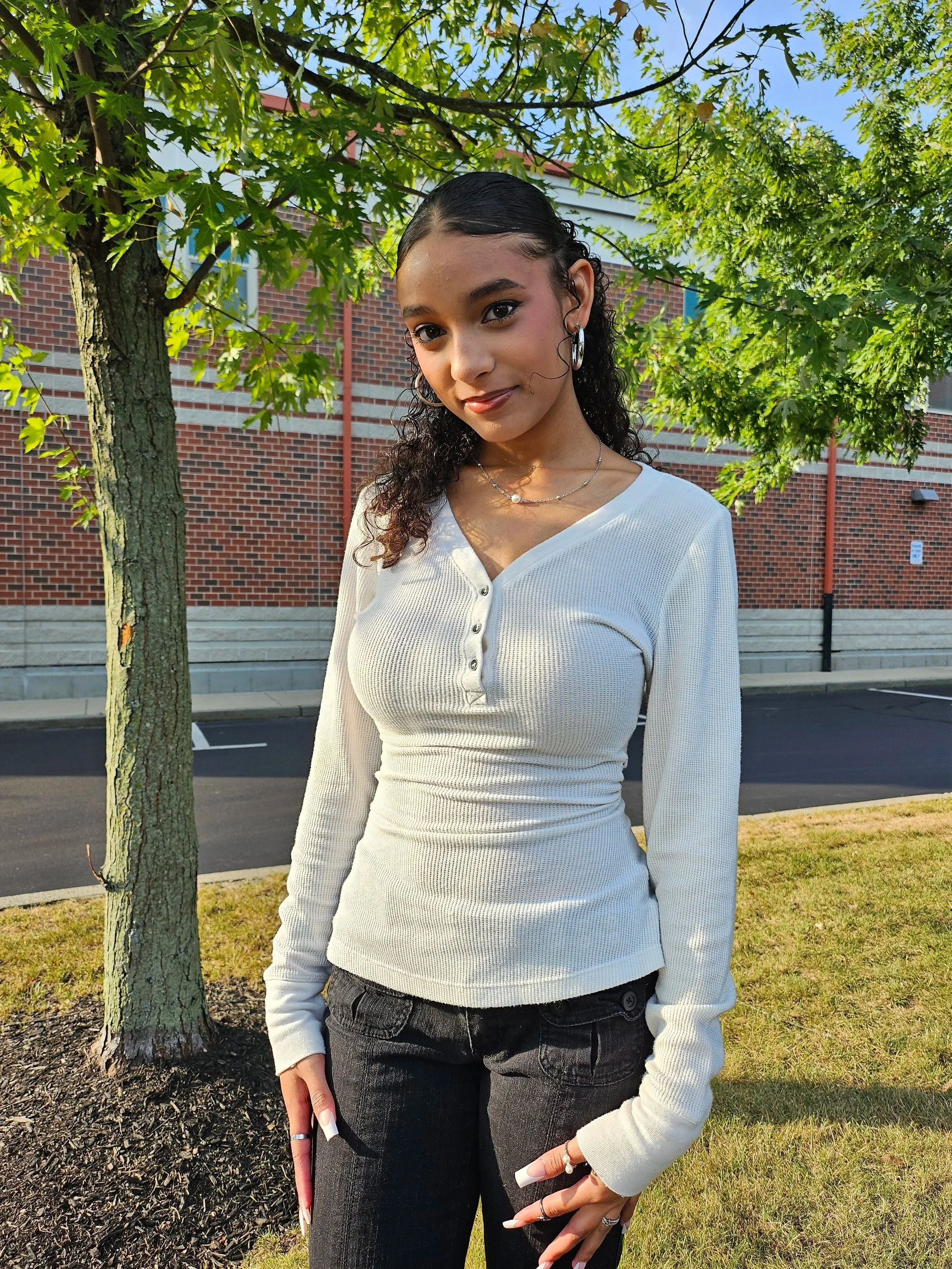 A young woman standing outdoors under a tree with green leaves, wearing a white long-sleeve top and black jeans, with jewelry including earrings and necklaces, and making a peace sign gesture with her right hand.