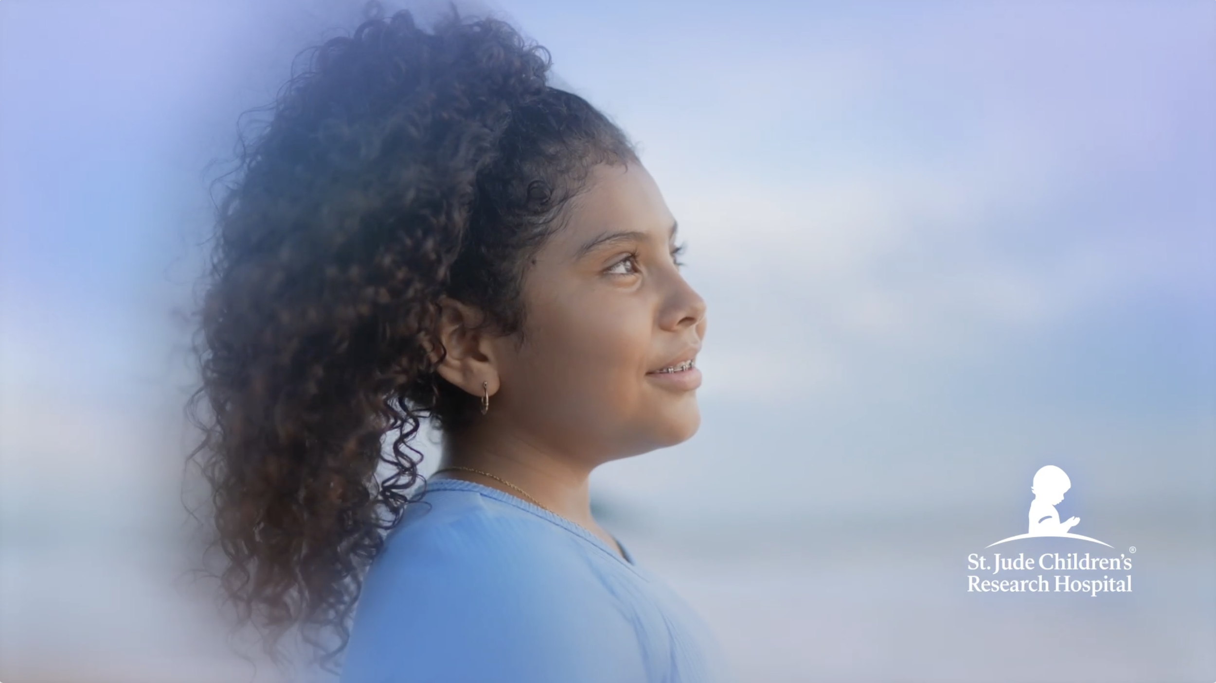 A young girl with curly hair looking towards the ocean during sunset, smiling softly, wearing a light blue shirt and gold jewelry, with a logo for St. Jude Children's Research Hospital in the bottom right corner.