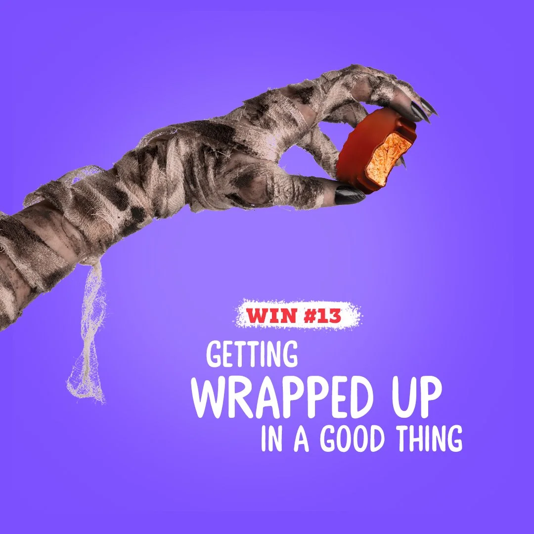 A zombie-like hand with black nails holding a red Reese's Peanut Butter Cup against a purple background. Text: 'Win #13 Getting Wrapped Up in a Good Thing.'