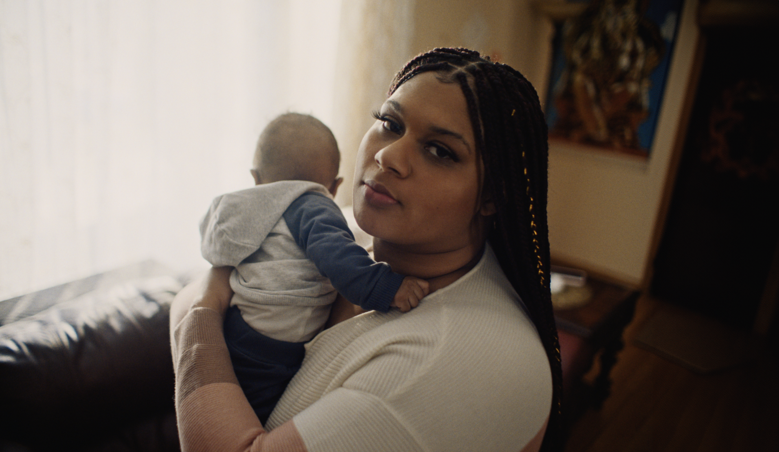 A woman with braided hair holding a young child, standing indoors near a window.