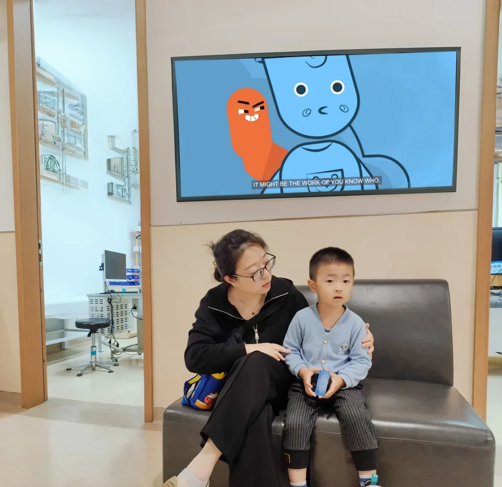 A woman and a young boy sitting on a bench in a medical facility waiting area. The woman is looking at the boy and has her arm around him, while the boy is holding a smartphone. There is a large screen above them displaying animated characters with subtitles.
