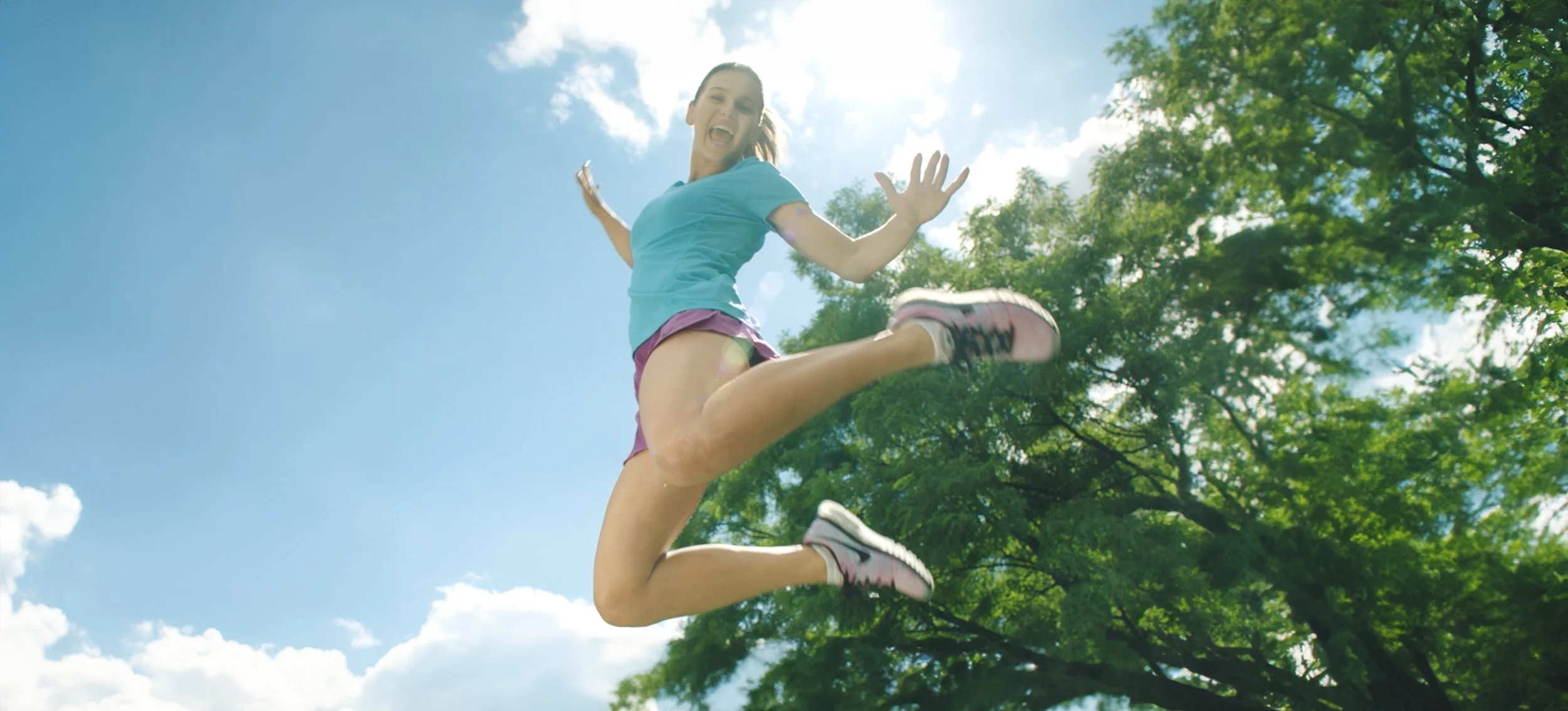 Young girl jumping in the air outdoors under a bright blue sky with clouds and green trees, wearing a light blue T-shirt, purple shorts, and pink and black sneakers.