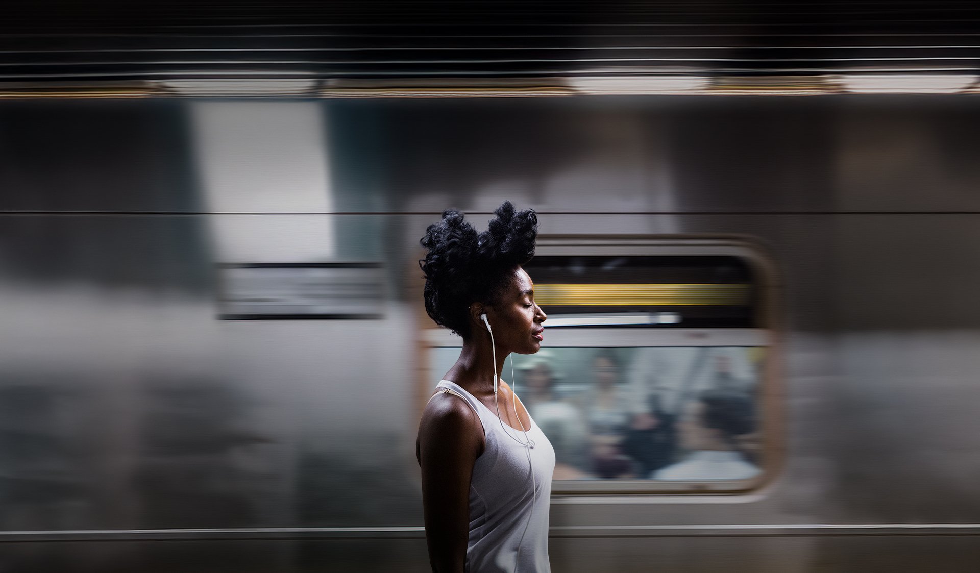 Woman with curly hair wearing a tank top and earphones walking past a moving train.