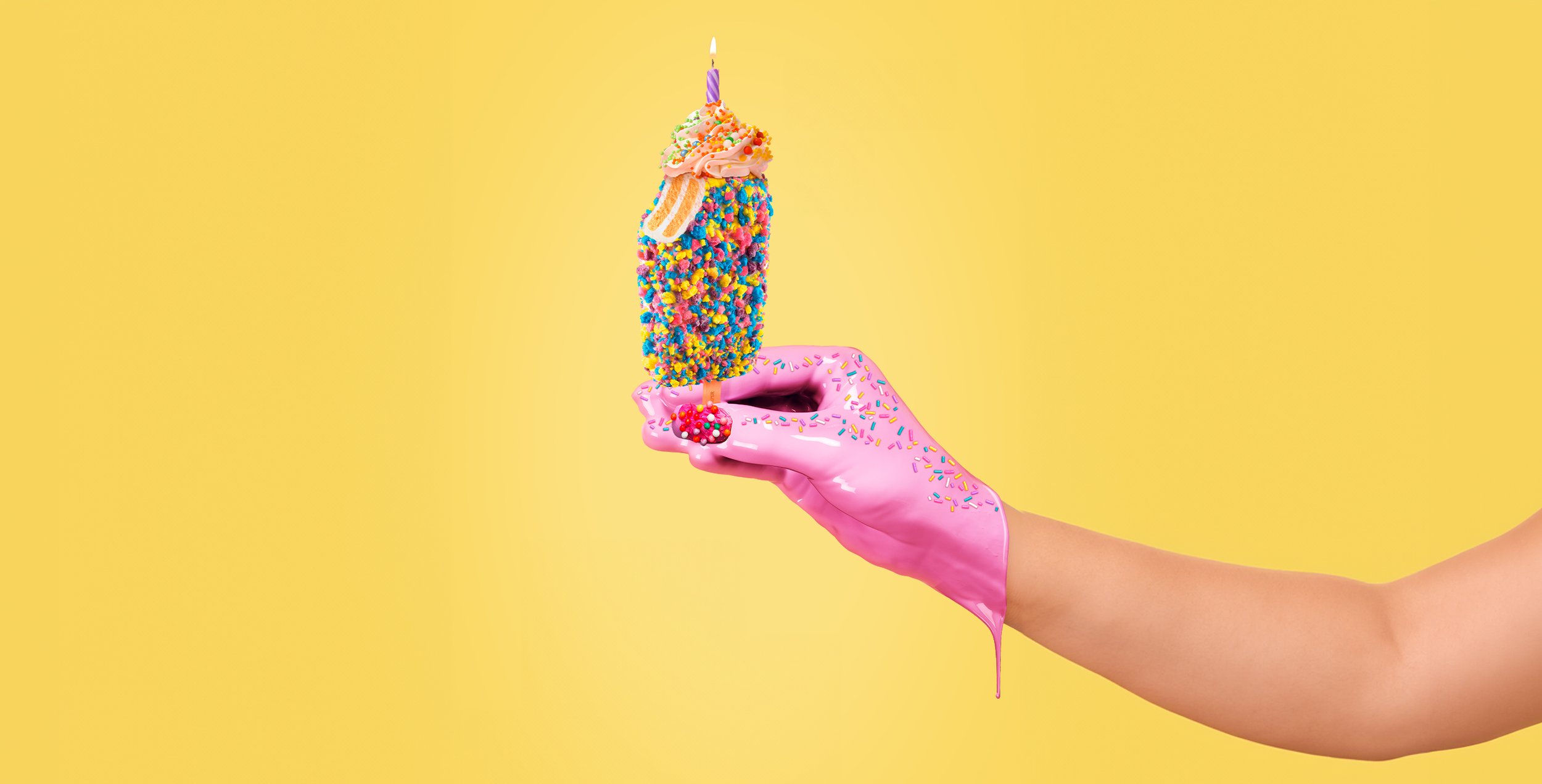 A hand wearing a pink, sparkly, sprinkle-covered glove holds a colorful, decorated birthday popicle with a birthday candle on top against a yellow background.