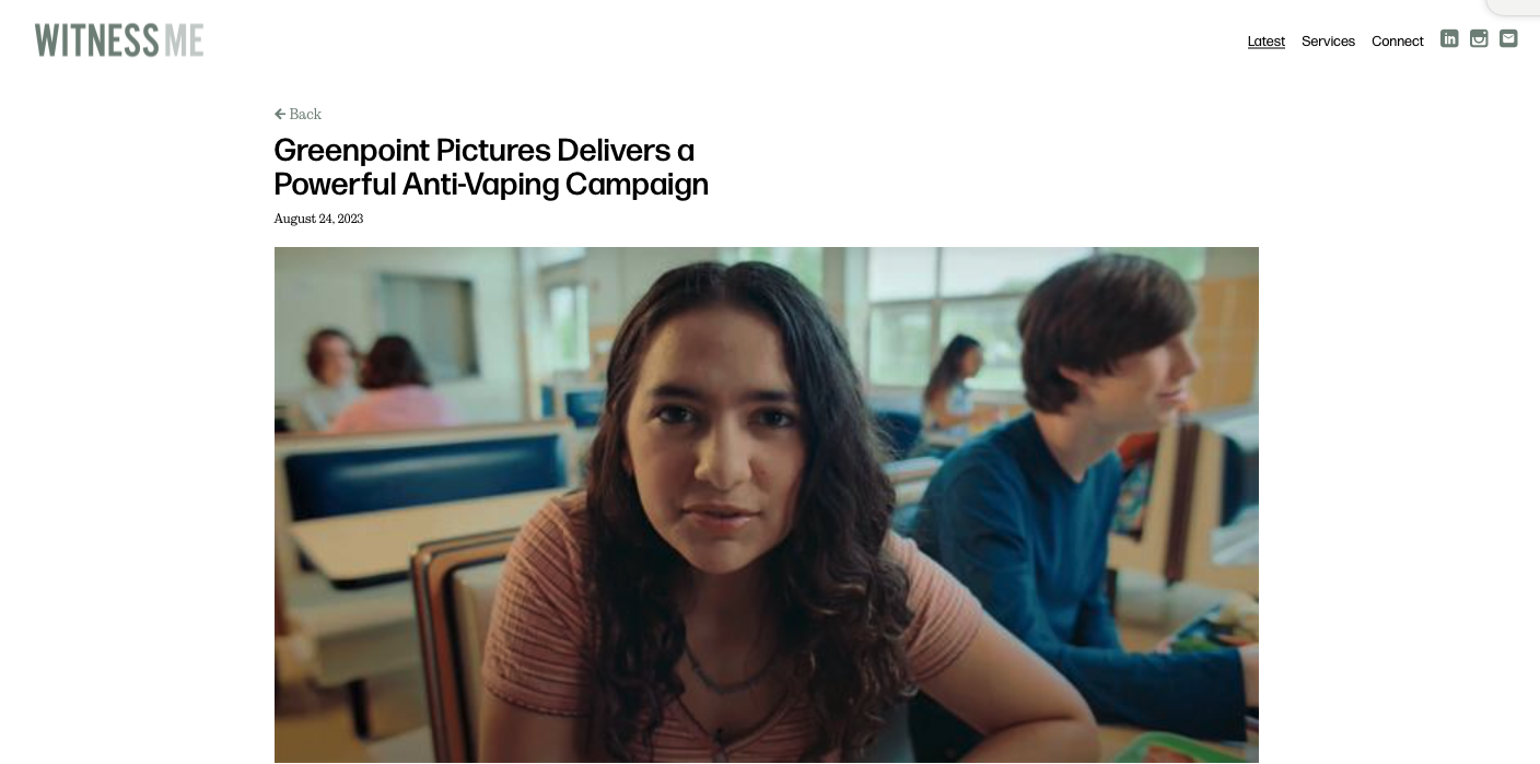 A young woman with long, dark hair sitting at a school desk, facing the camera, with other students in the background.