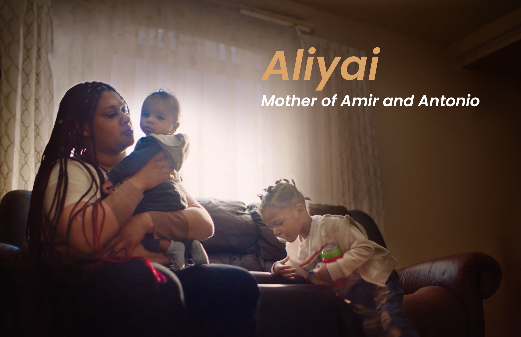 A woman holding a baby on a sofa with a boy sitting on the floor looking at a tablet in a sunlit living room.