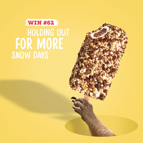 A hand holding a large chocolate-covered ice cream bar with a nut topping, casting a shadow on a yellow background. Text reads 'Win #62 Holding out for more snow days.'