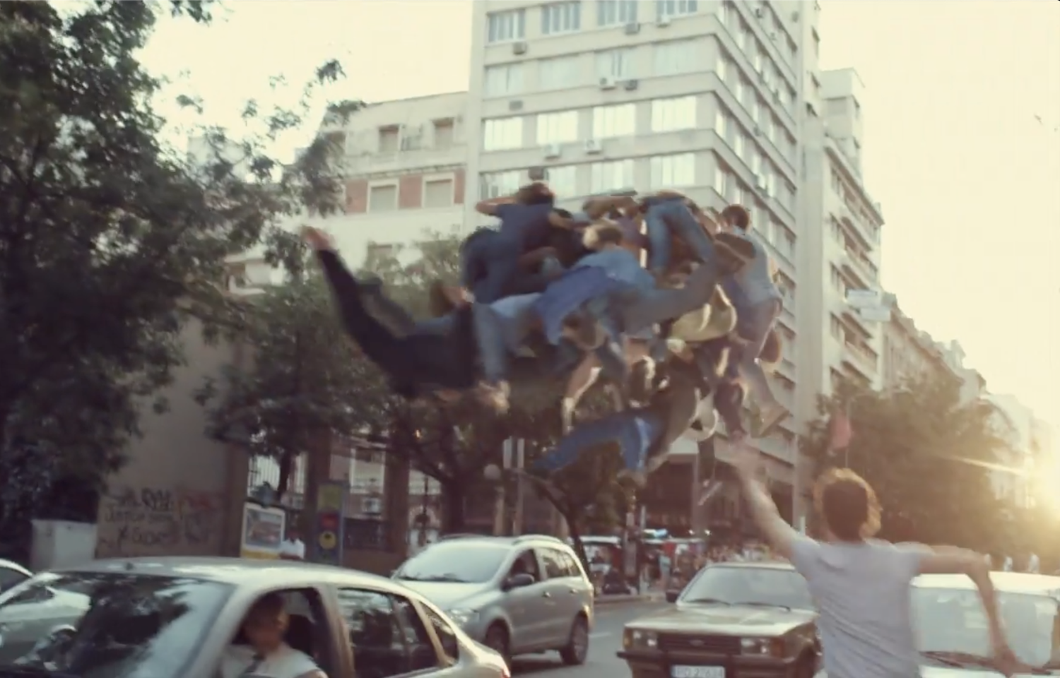 A group of people stacked on top of each other in the air above parked cars on a city street during sunset.