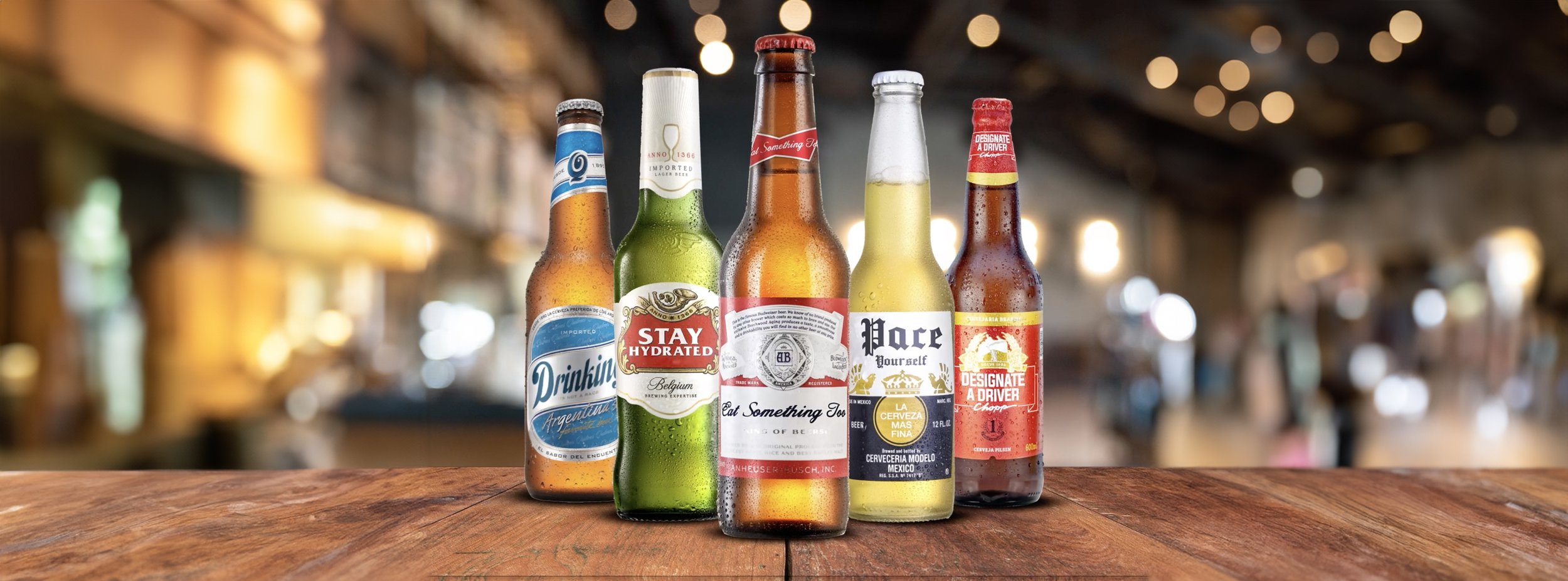 Six different bottled beers arranged on a wooden surface in a bar setting, with blurred bar background and warm lighting.