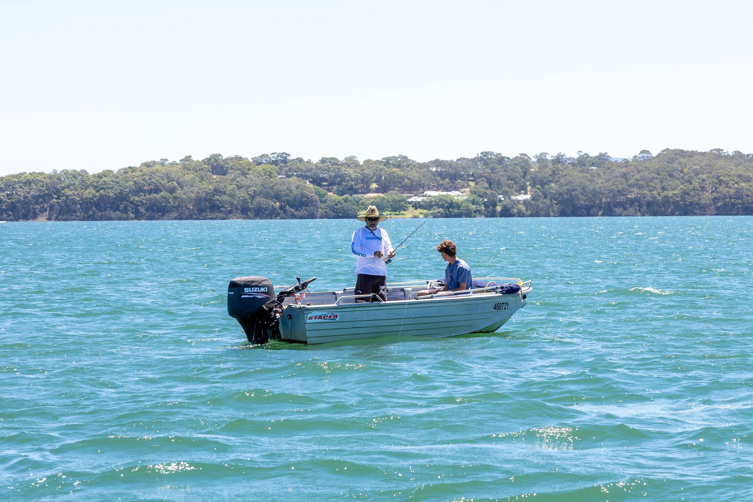 4.5m boat hire - Lake Macquarie.Two people fishing on a small boat in a body of water with a tree-lined shore in the background.