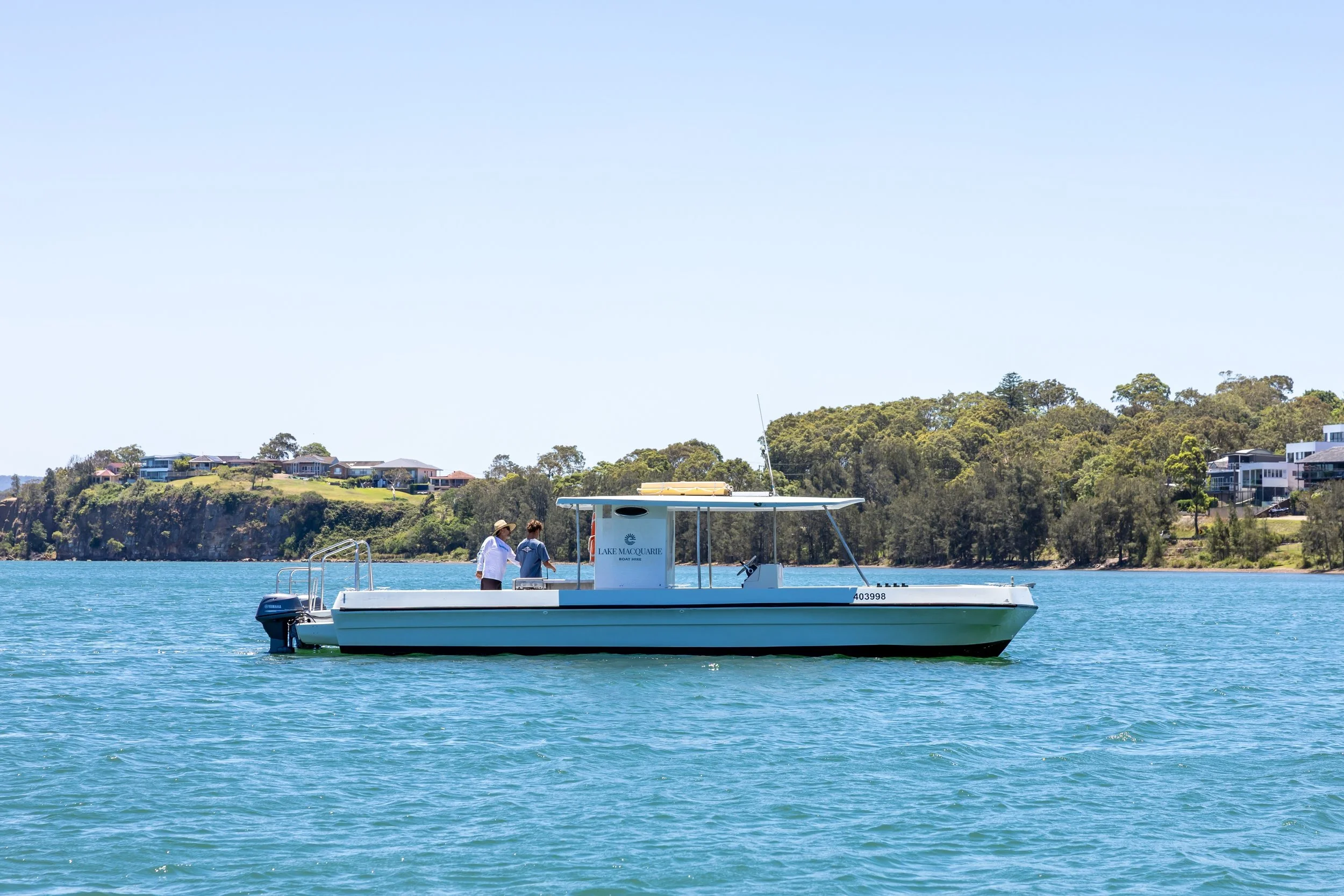 Day Tripper BBQ Pontoon Boat - Lake Macquarie. A boat with two people on a lake with houses and trees in the background.