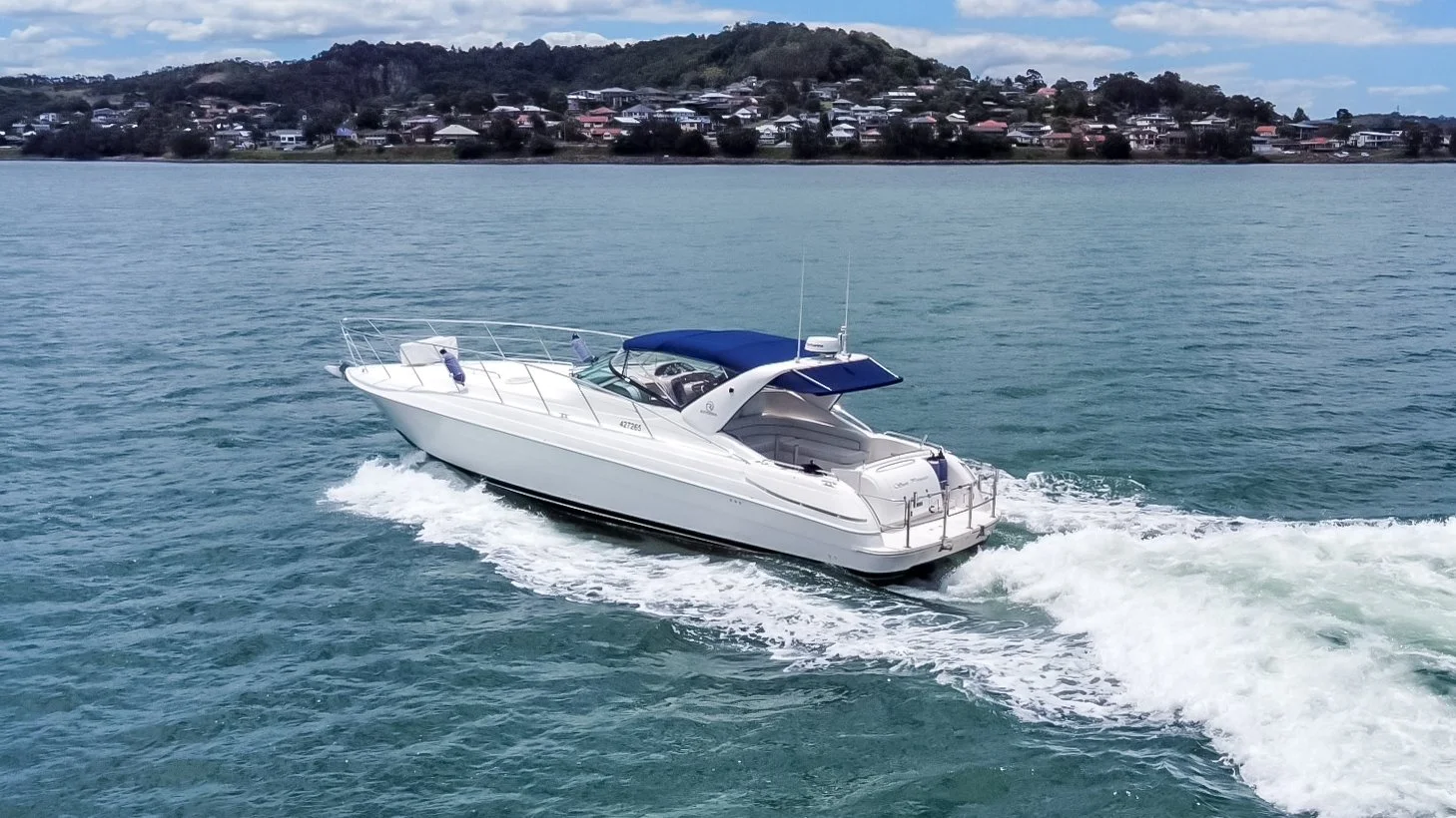 A white motor yacht with a blue canopy cruising on a body of water, with a residential shoreline and houses visible in the background.