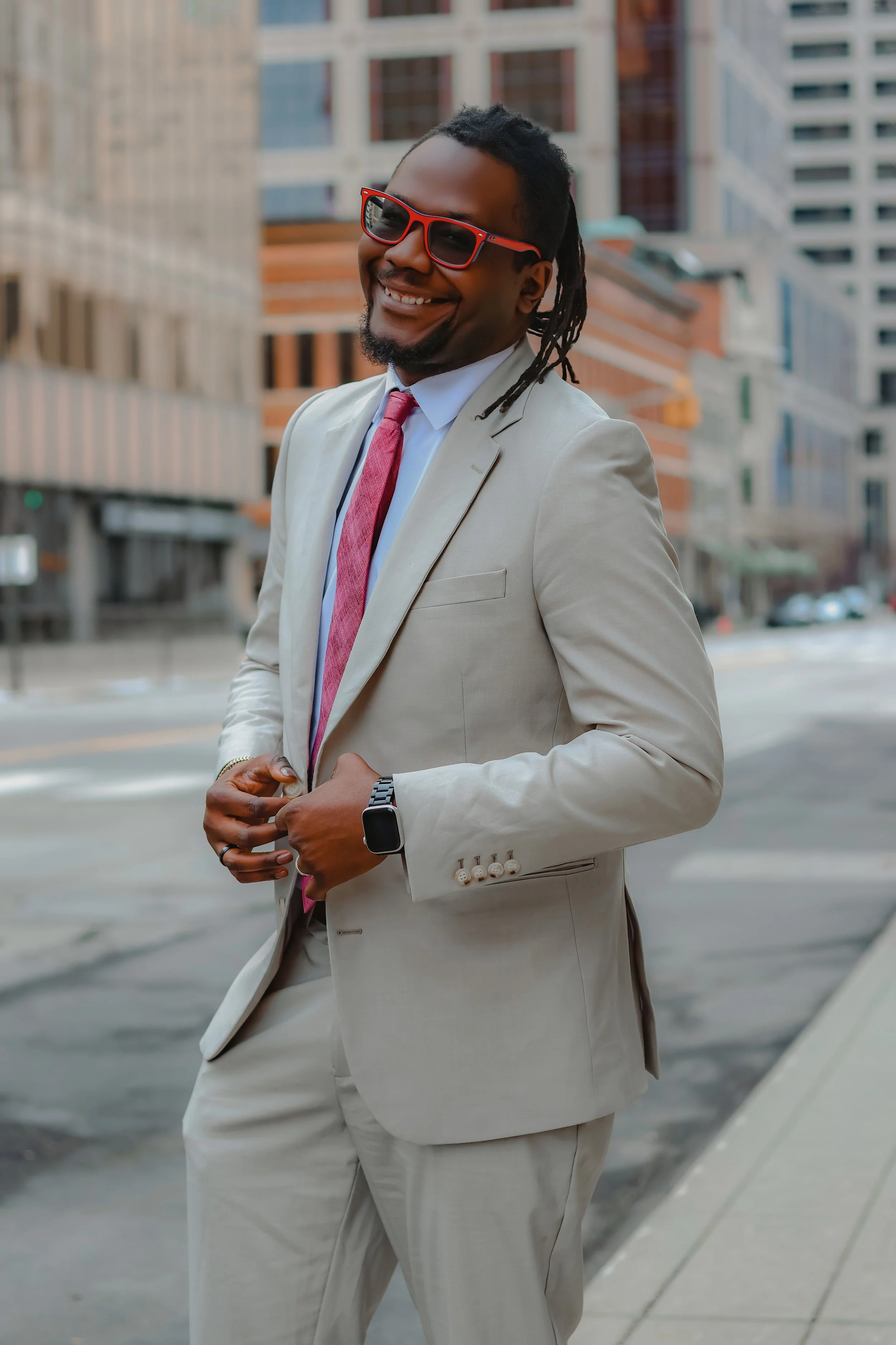 A man in a white suit, pink tie, and sunglasses smiling outdoors in an urban setting with tall buildings in the background.