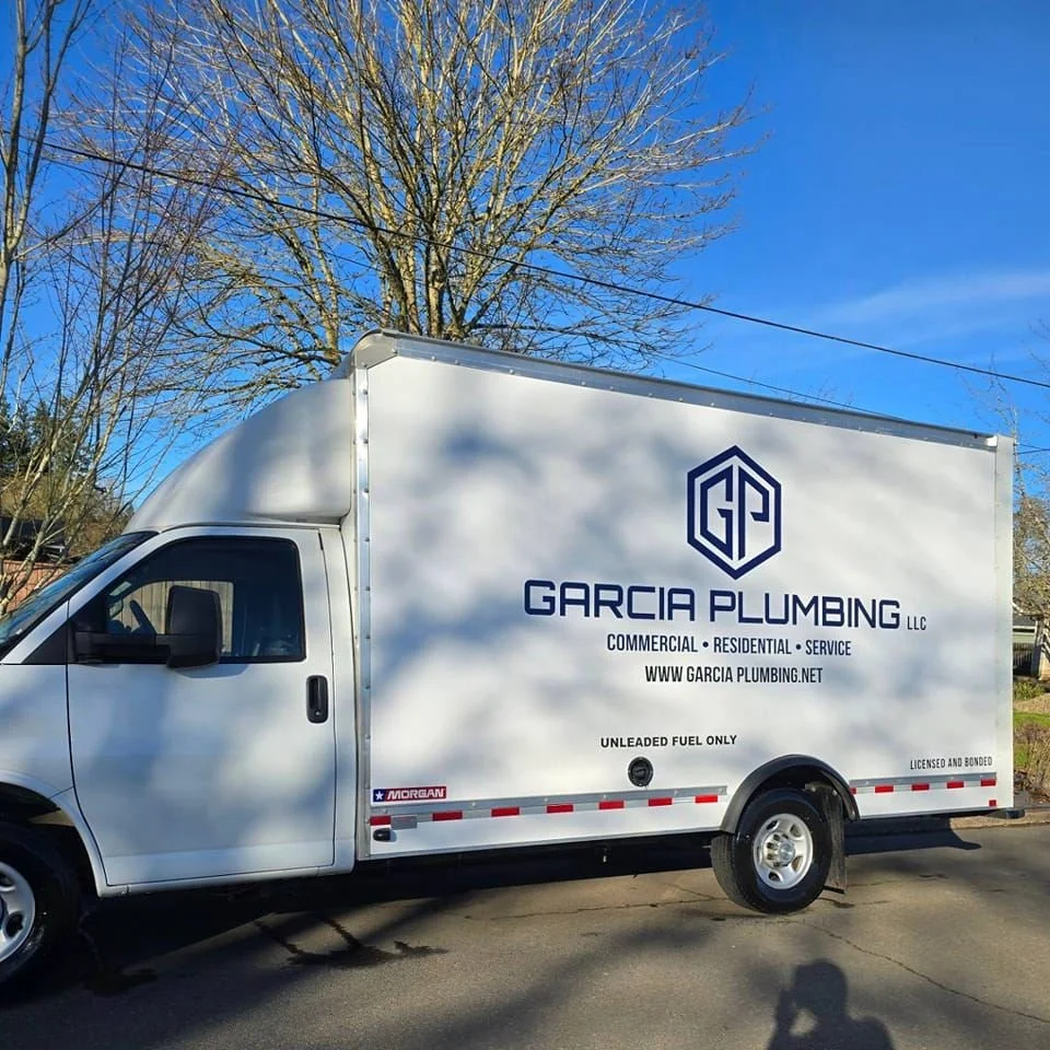 A white service truck with Garcia Plumbing LLC logo and text, parked on a street with leafless trees and a clear blue sky in the background.