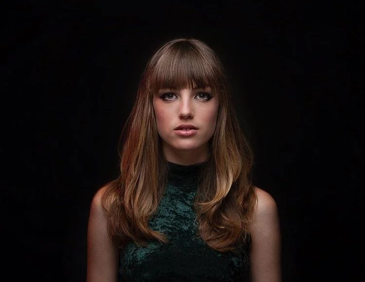 Portrait of a young woman with brown hair and bangs, wearing a dark sleeveless top, looking directly at the camera against a black background.